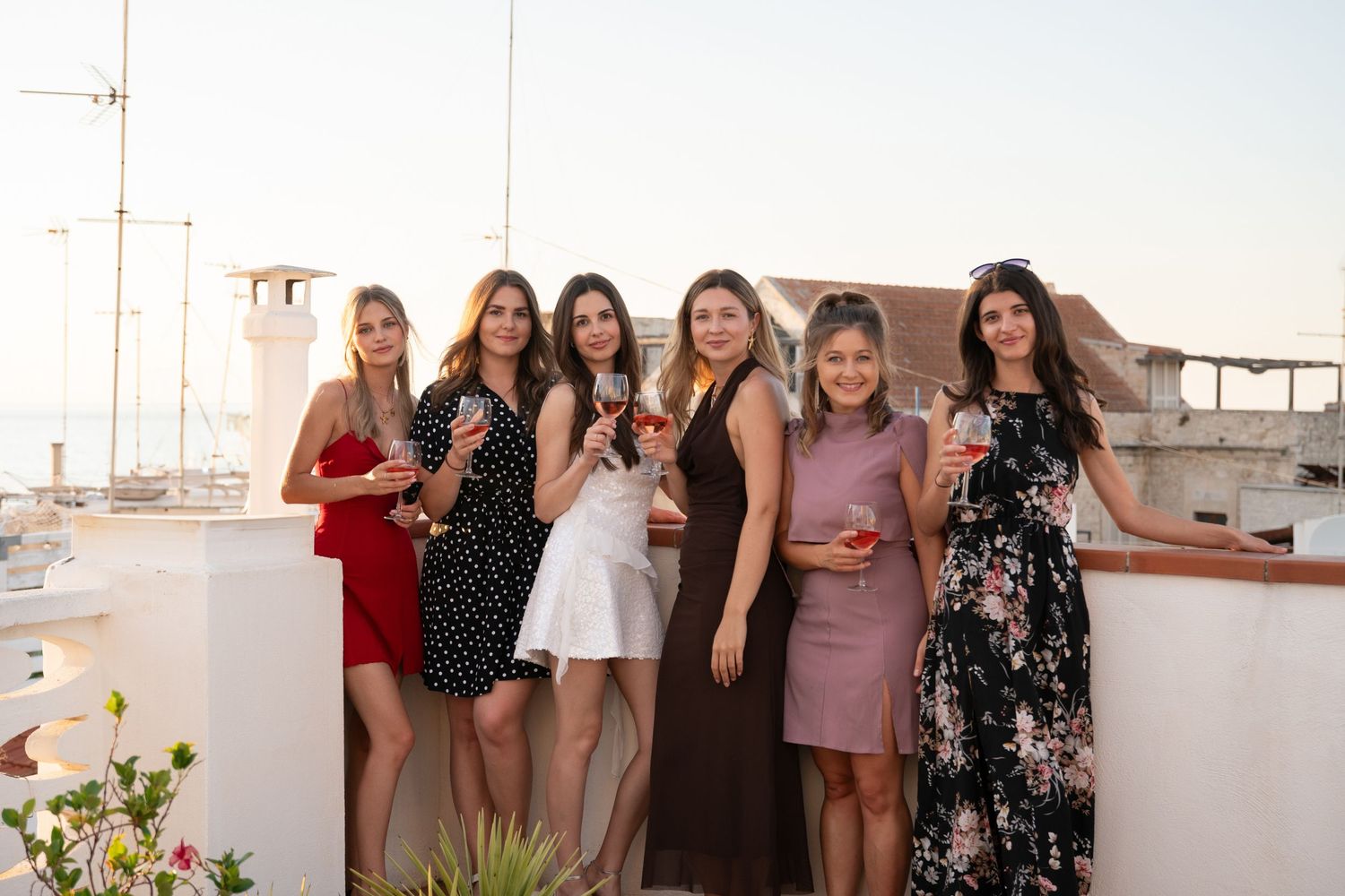 A group of women in cocktail dresses posing together on a rooftop terrace at sunset.