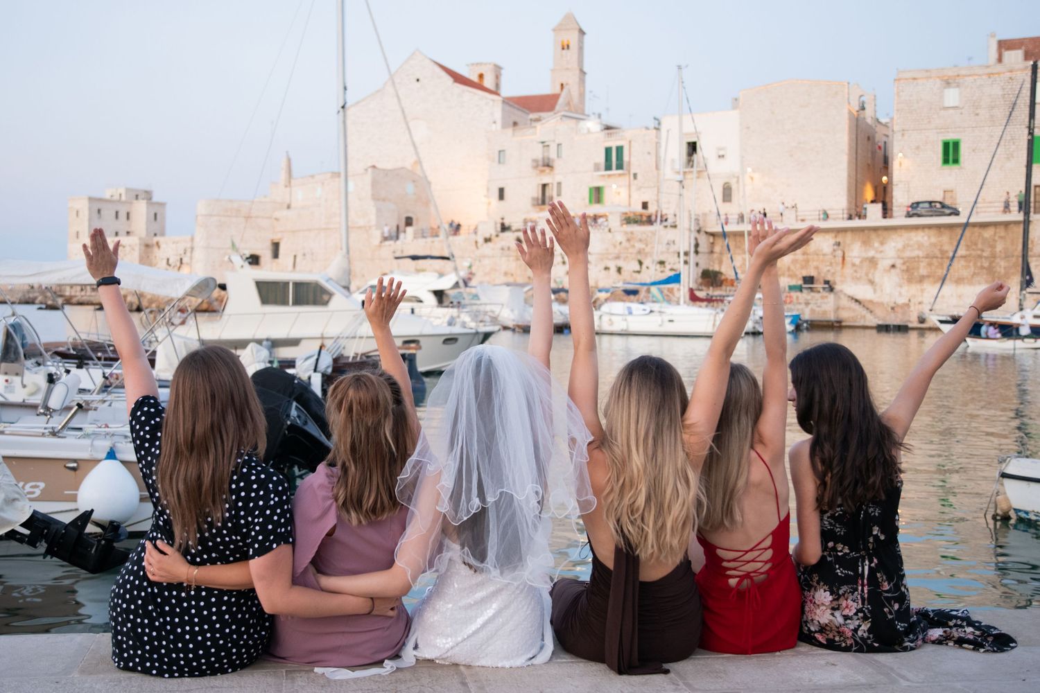Group of friends sitting on seaside wall overlooking harbor with boats and buildings in background.