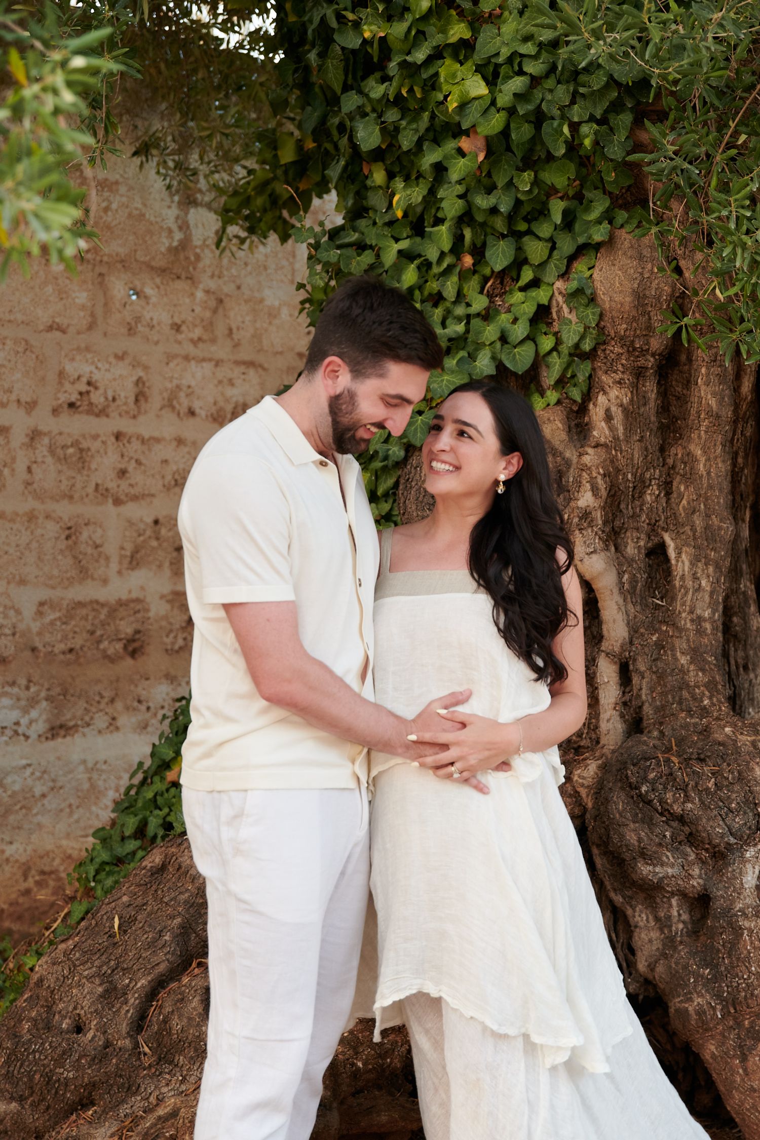 A romantic couple in white attire posing against an ivy-covered wall.