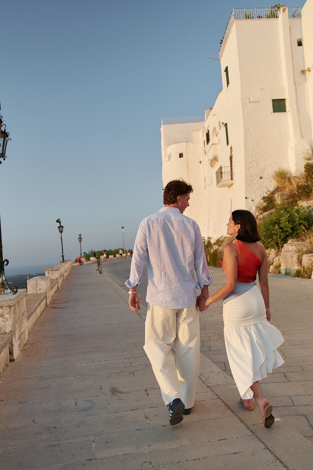 Couple walking along a white Mediterranean pathway at sunset.