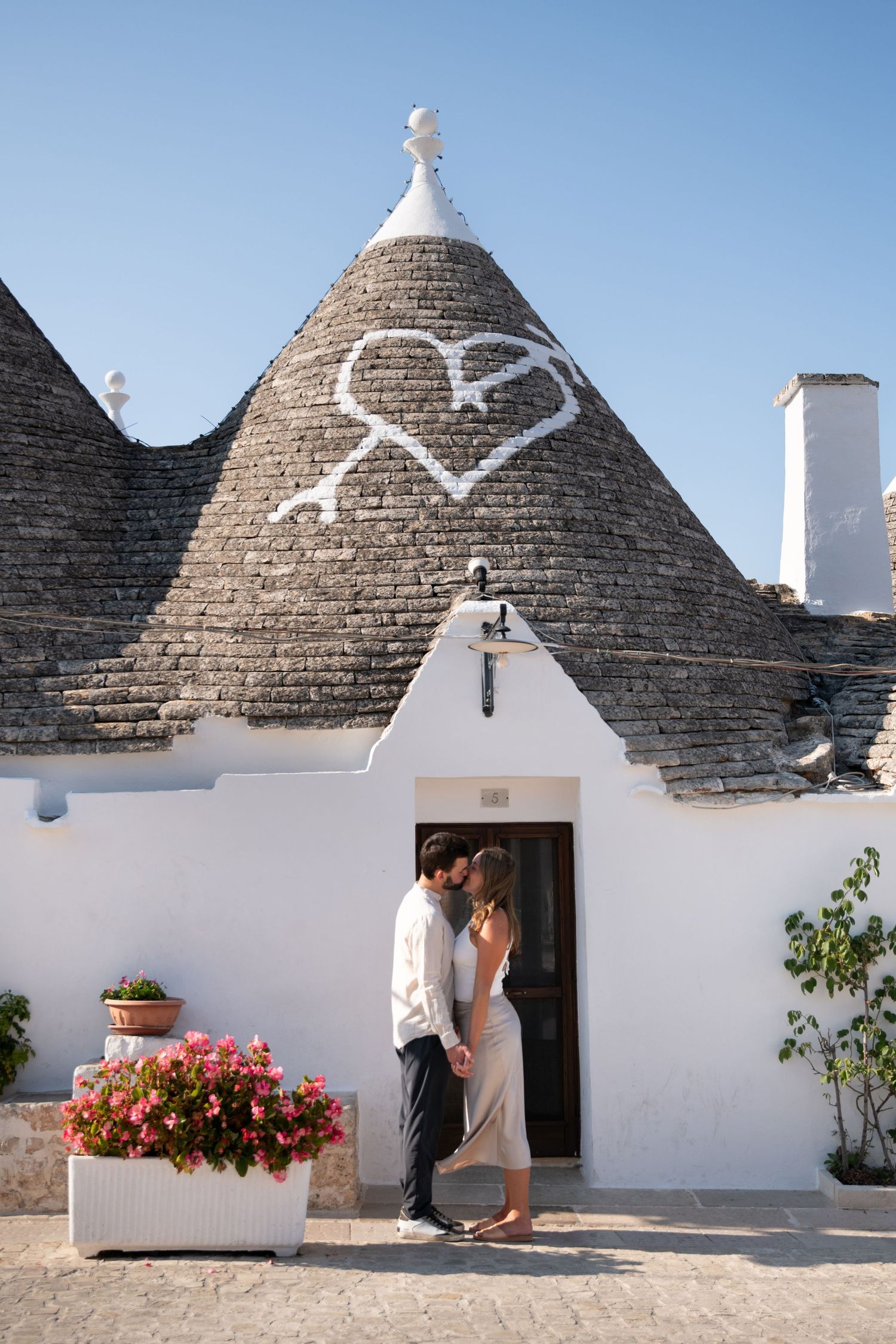 Romantic moment captured at traditional white trulli buildings with conical roofs and pink flowers.