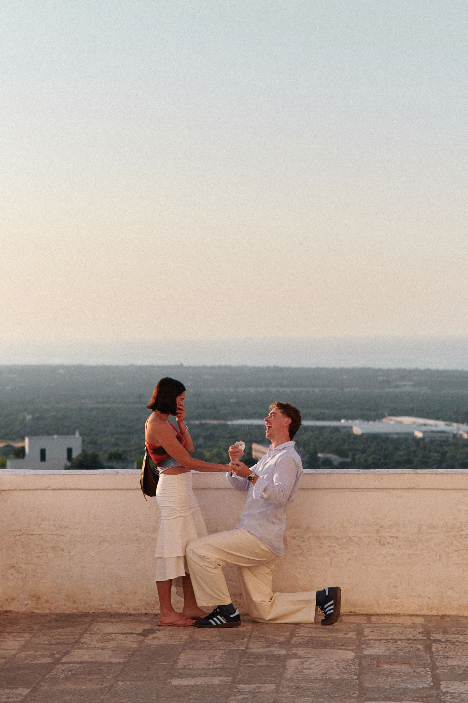 A romantic proposal moment at sunset on a scenic overlook with city views in the background.