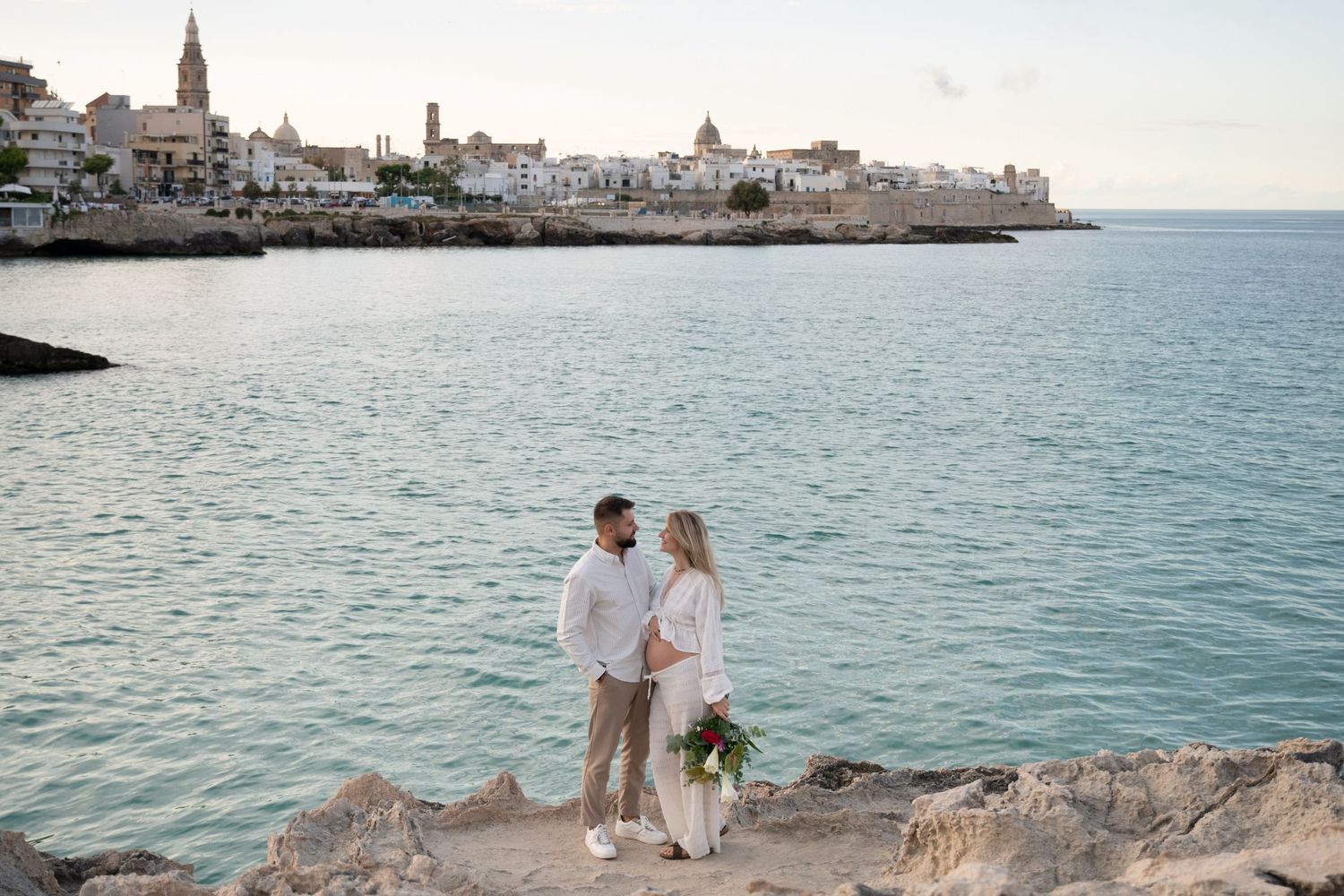 Couple in white clothing walking along rocky coastal edge with seaside town view.