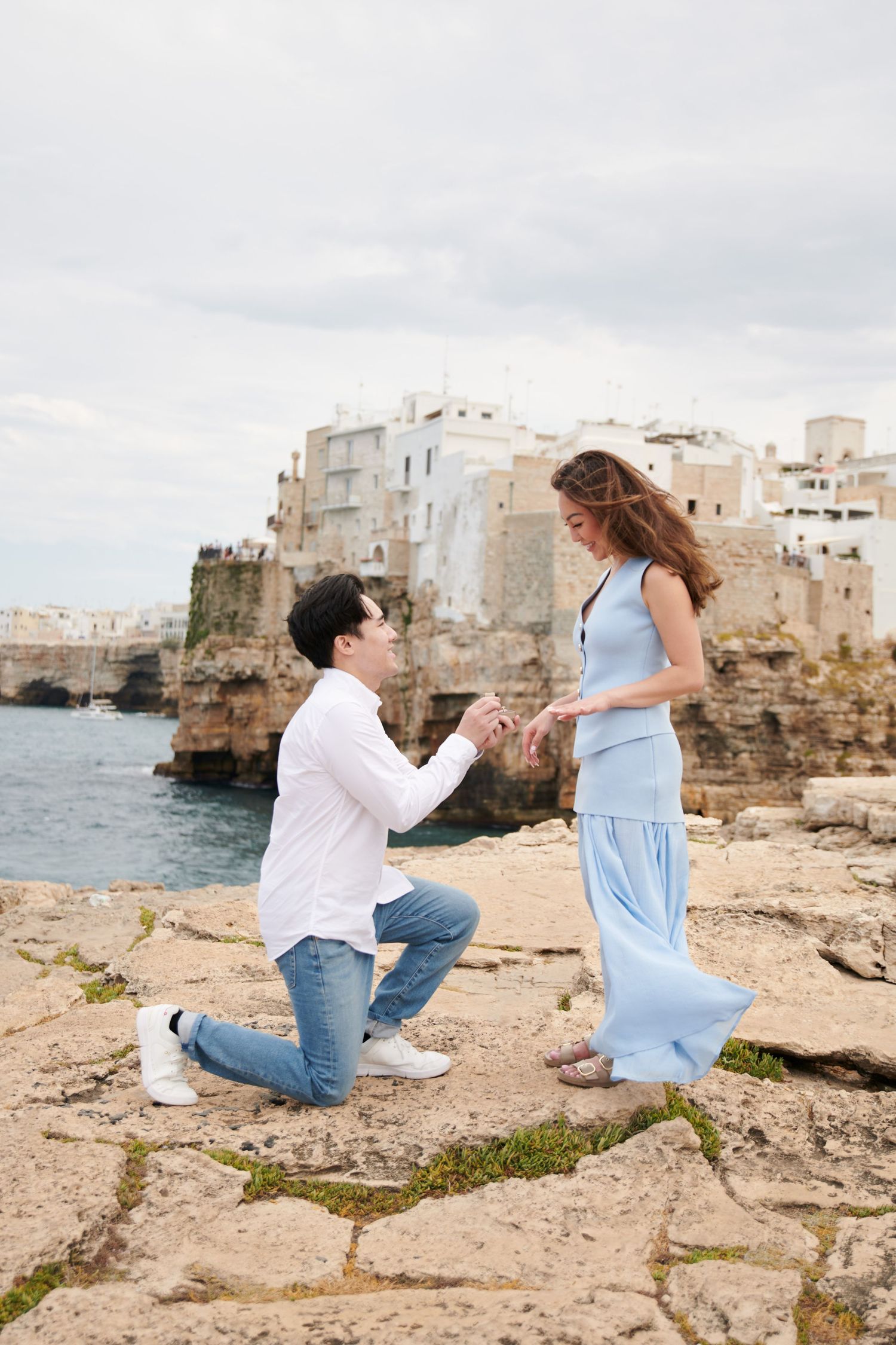 Surprise proposal moment captured on rocky coastal cliff with Mediterranean town in background.