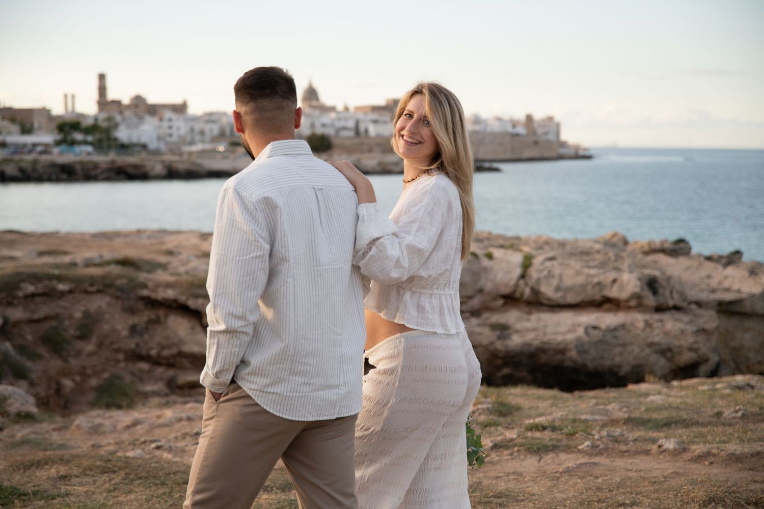 Romantic couple embraces on a rocky coastline overlooking a Mediterranean seaside town at sunset.