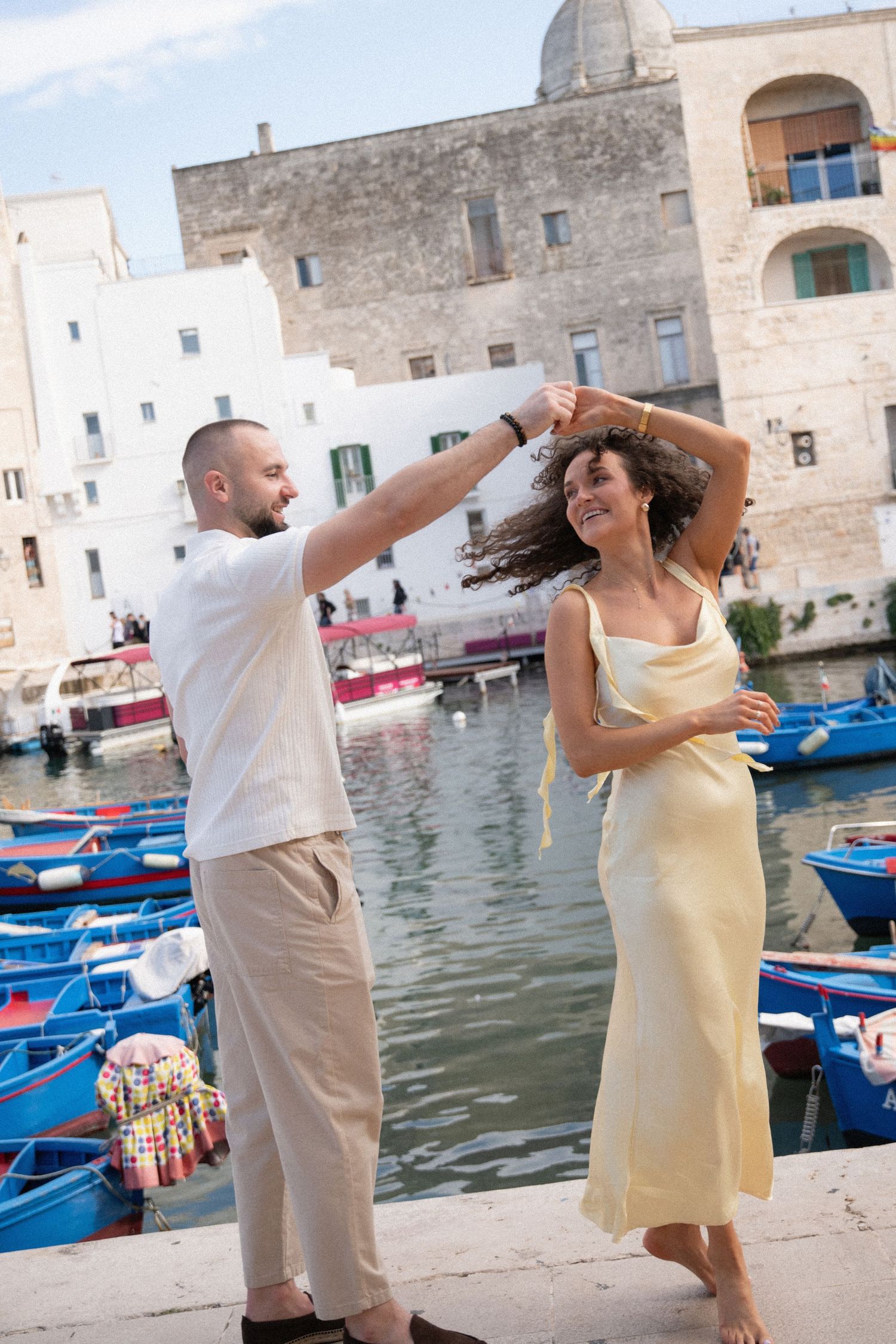 Couple in yellow dress and white shirt dancing playfully by harbor with traditional boats.
