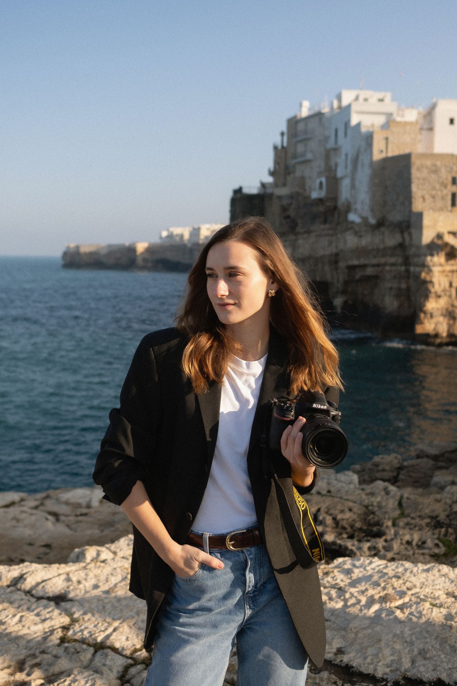 Photographer in black blazer stands on rocky coastline.