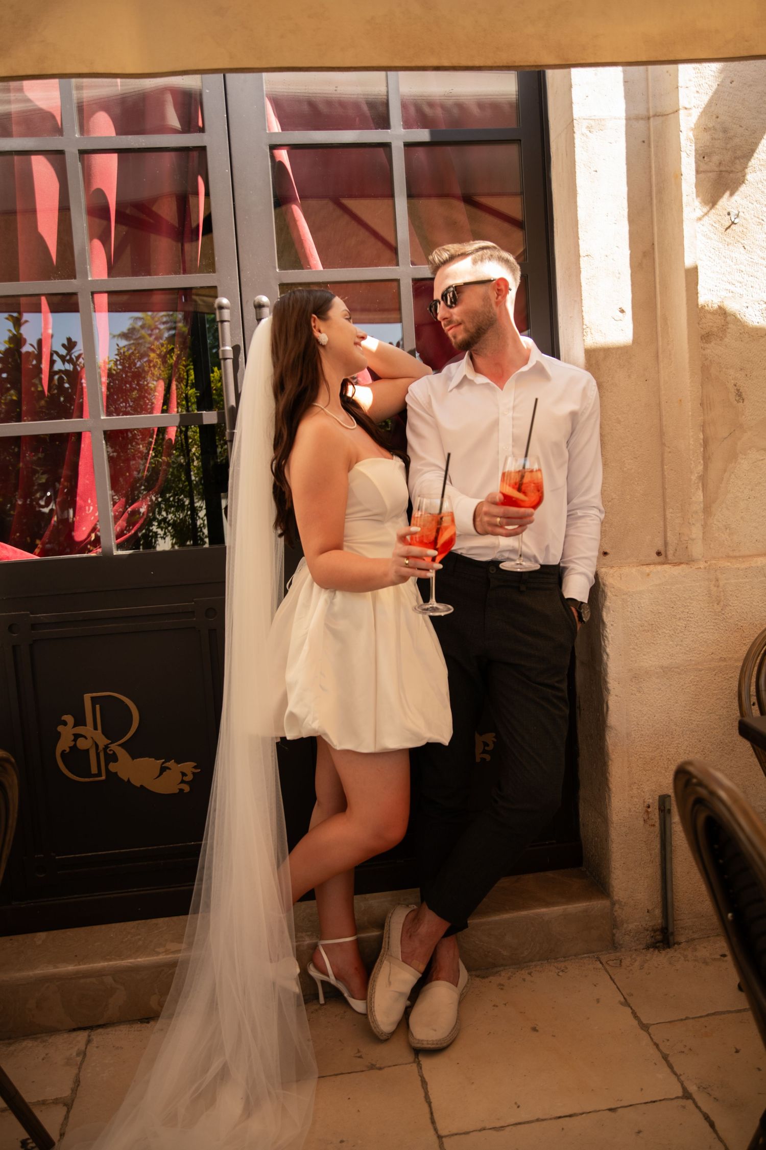 Couple in wedding attire shares drinks and intimate moment against rustic doorway.