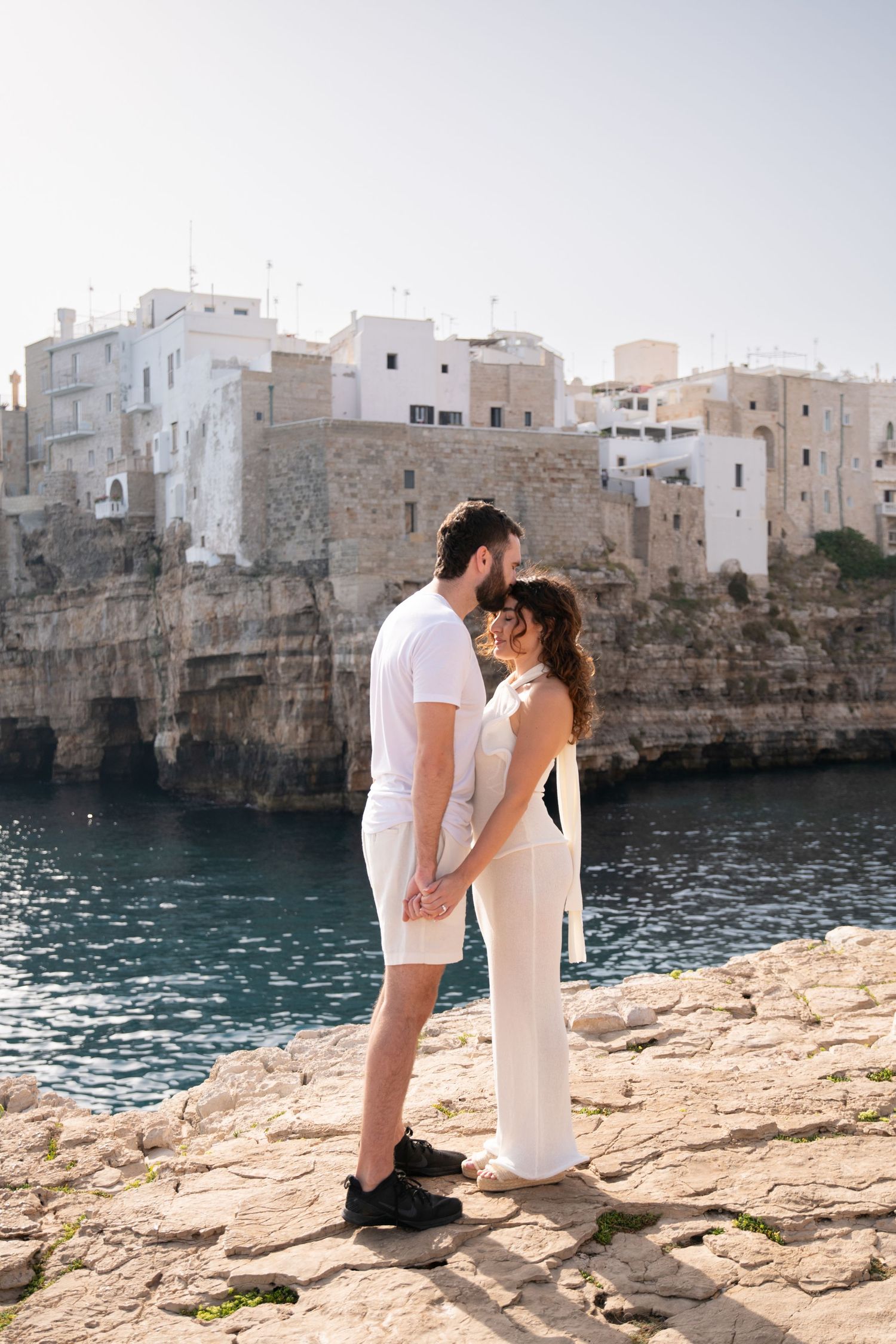 Couple stands on rocky coastal cliffs with white Mediterranean buildings and blue water in the background.