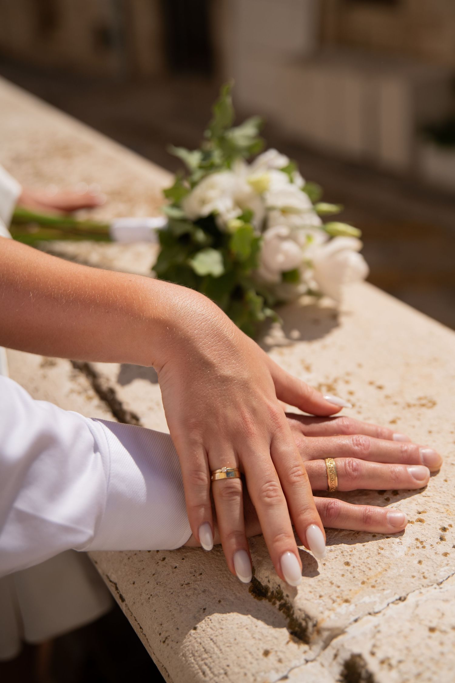Close-up of hands with wedding rings resting on a stone surface with white flowers.