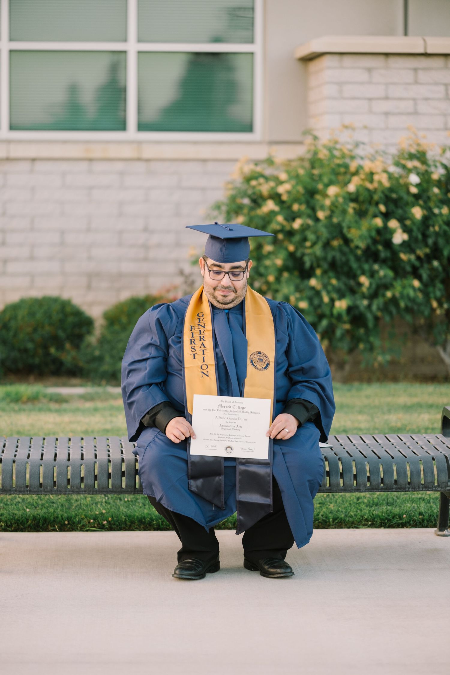 Graduate in blue cap and gown poses with diploma on campus bench.