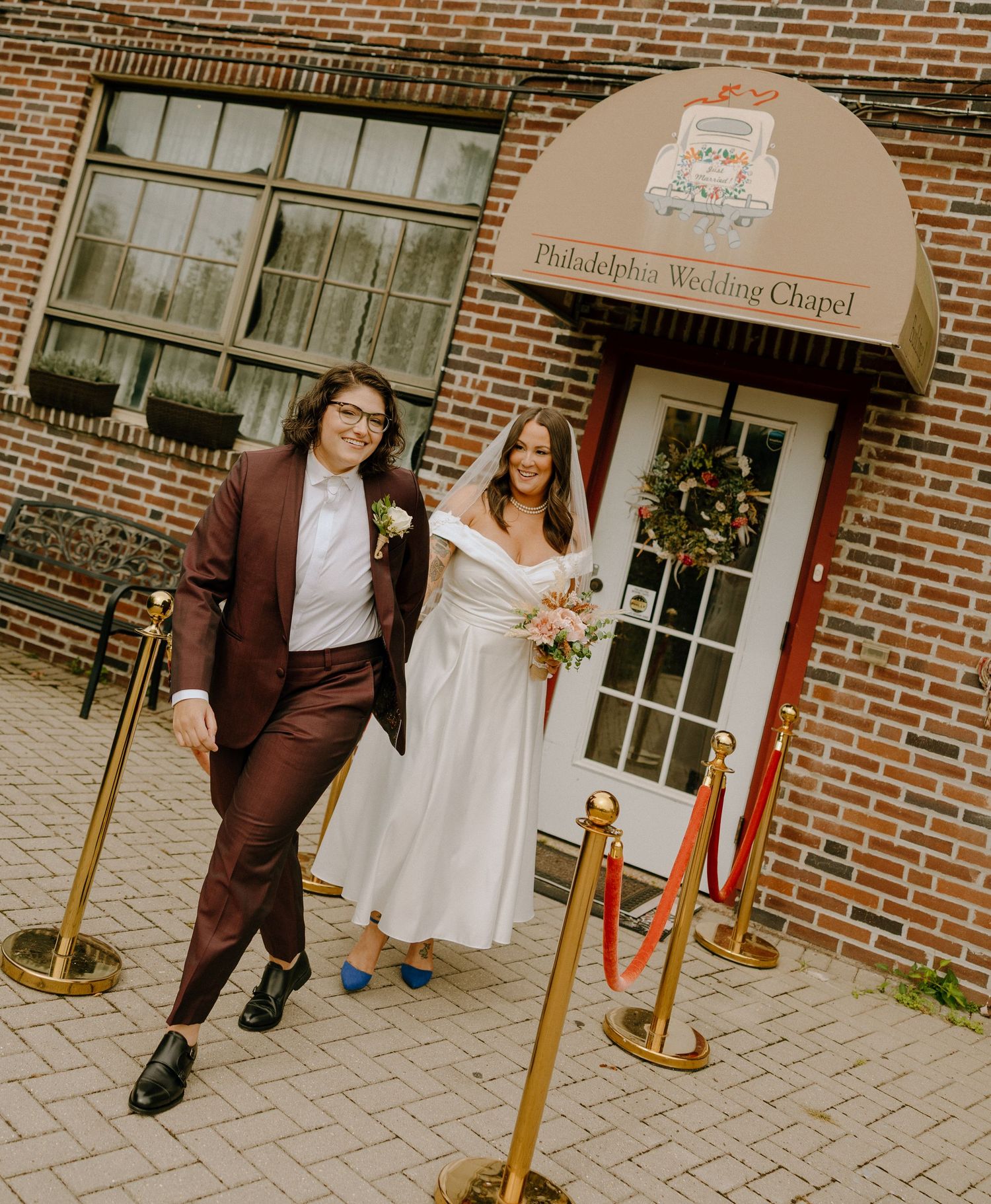 A couple poses outside a brick chapel entrance with a brown awning and decorative wreath on the door.
