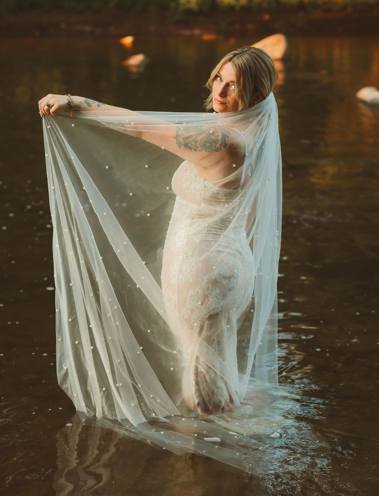 A bride in a fitted dress stands in water while dramatically holding up her flowing veil at sunset.