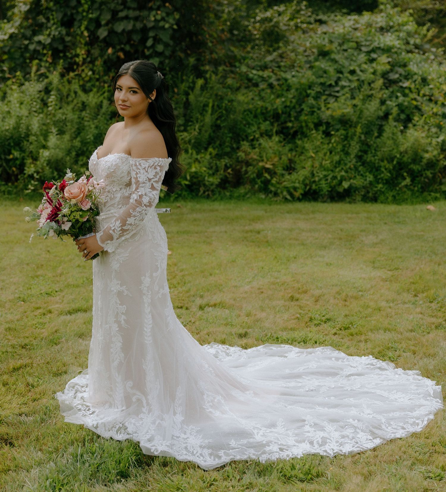 A flowing white lace wedding dress with intricate details trails across green grass in an outdoor setting.
