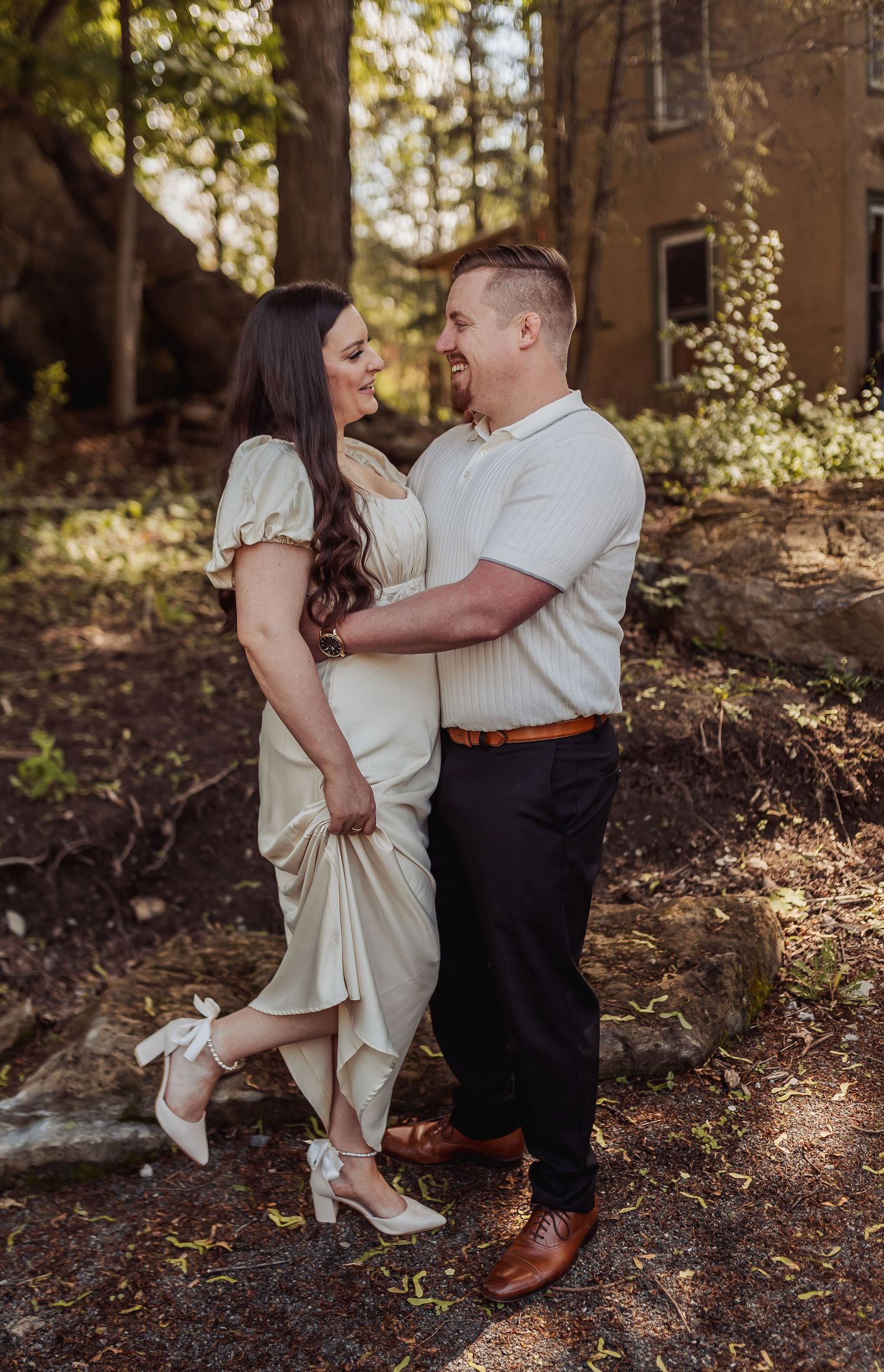 A couple poses together in dappled sunlight amongst trees and fallen leaves.