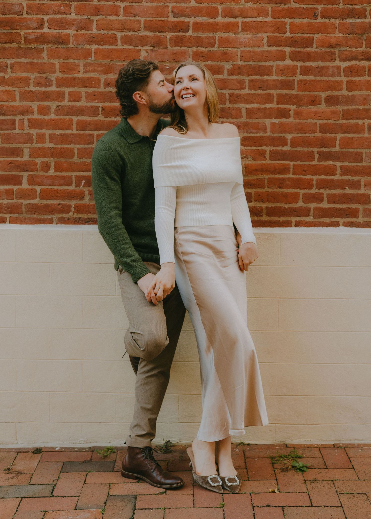 A couple shares a romantic moment against a rustic brick wall backdrop on a paved walkway.