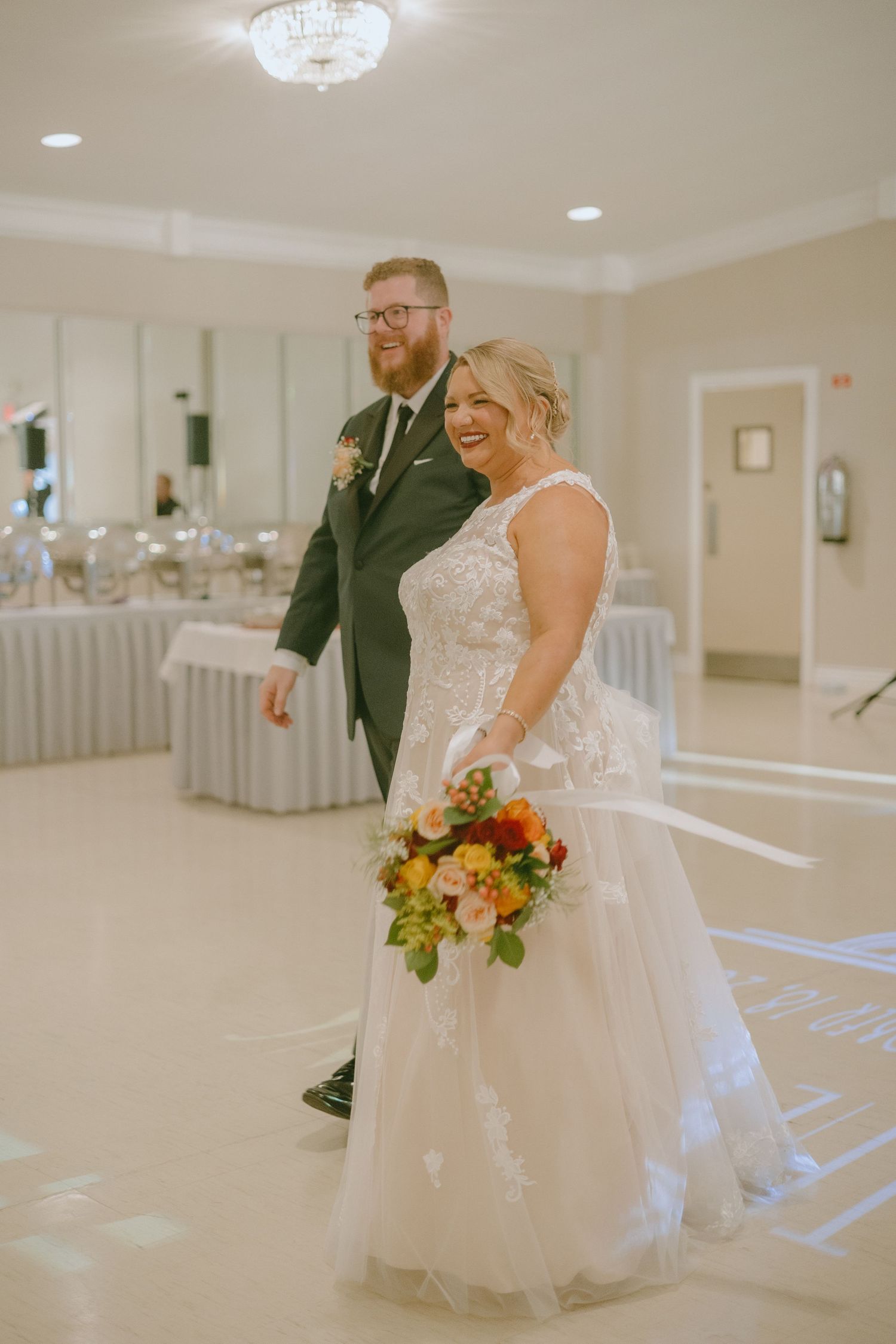 A couple in wedding attire dance together in an elegant white reception hall with soft lighting.