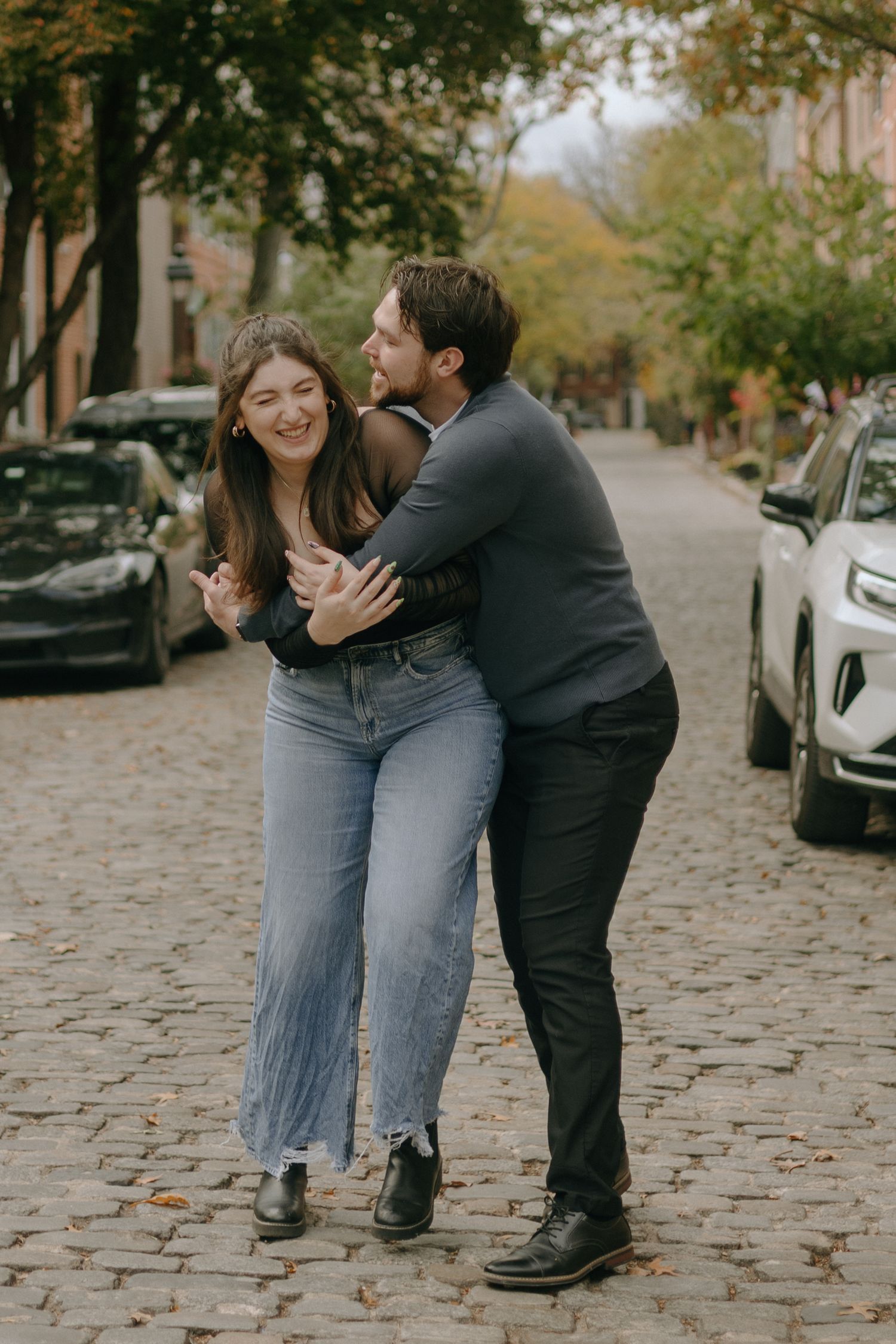 Couple playfully interacting on a cobblestone street lined with autumn trees and parked cars.