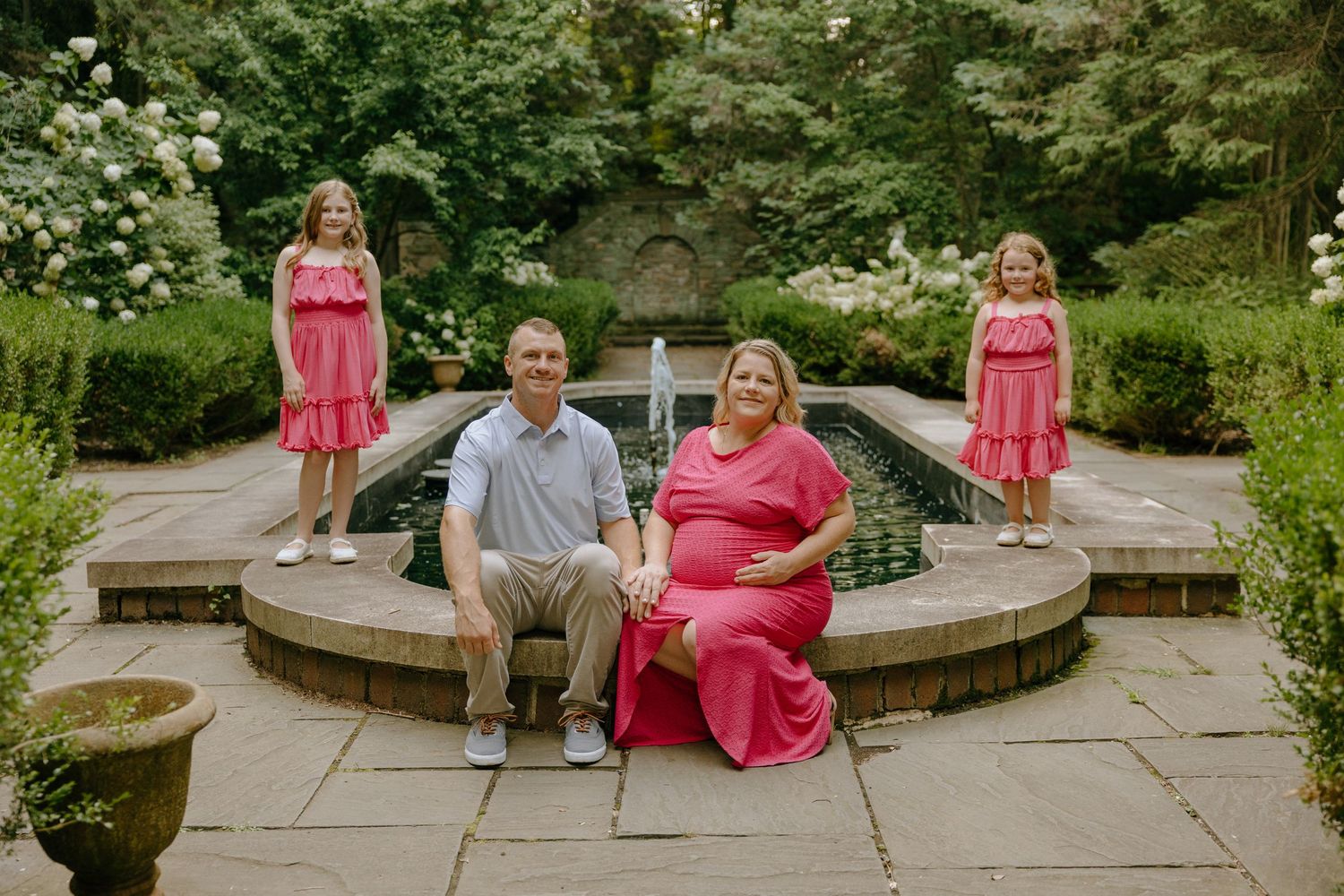 People in pink dresses sit on curved stone wall in lush garden setting.