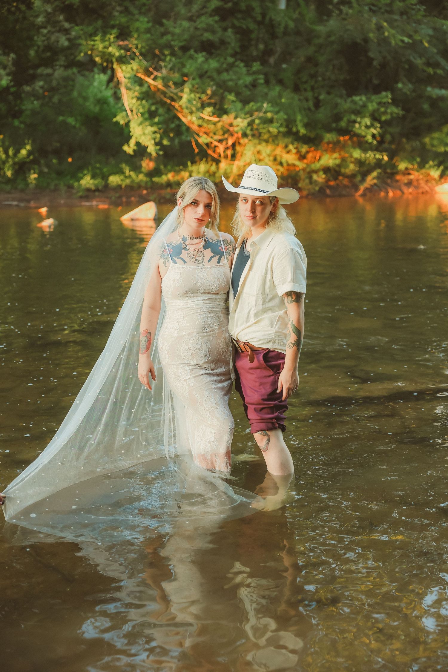A couple stands in a shallow river while wearing wedding attire and sharing a playful moment together.