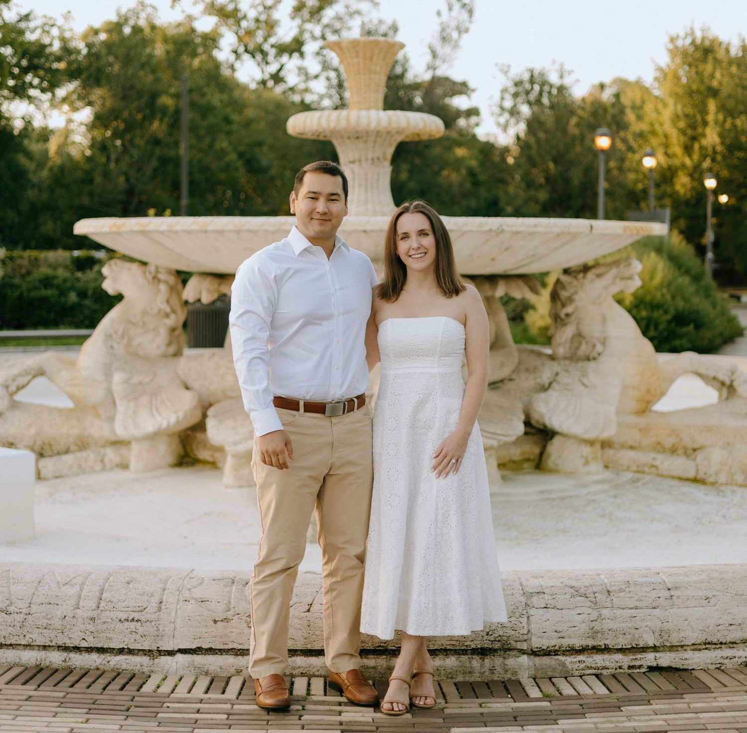 A couple poses in formal attire in front of an ornate stone fountain in a garden setting at sunset.