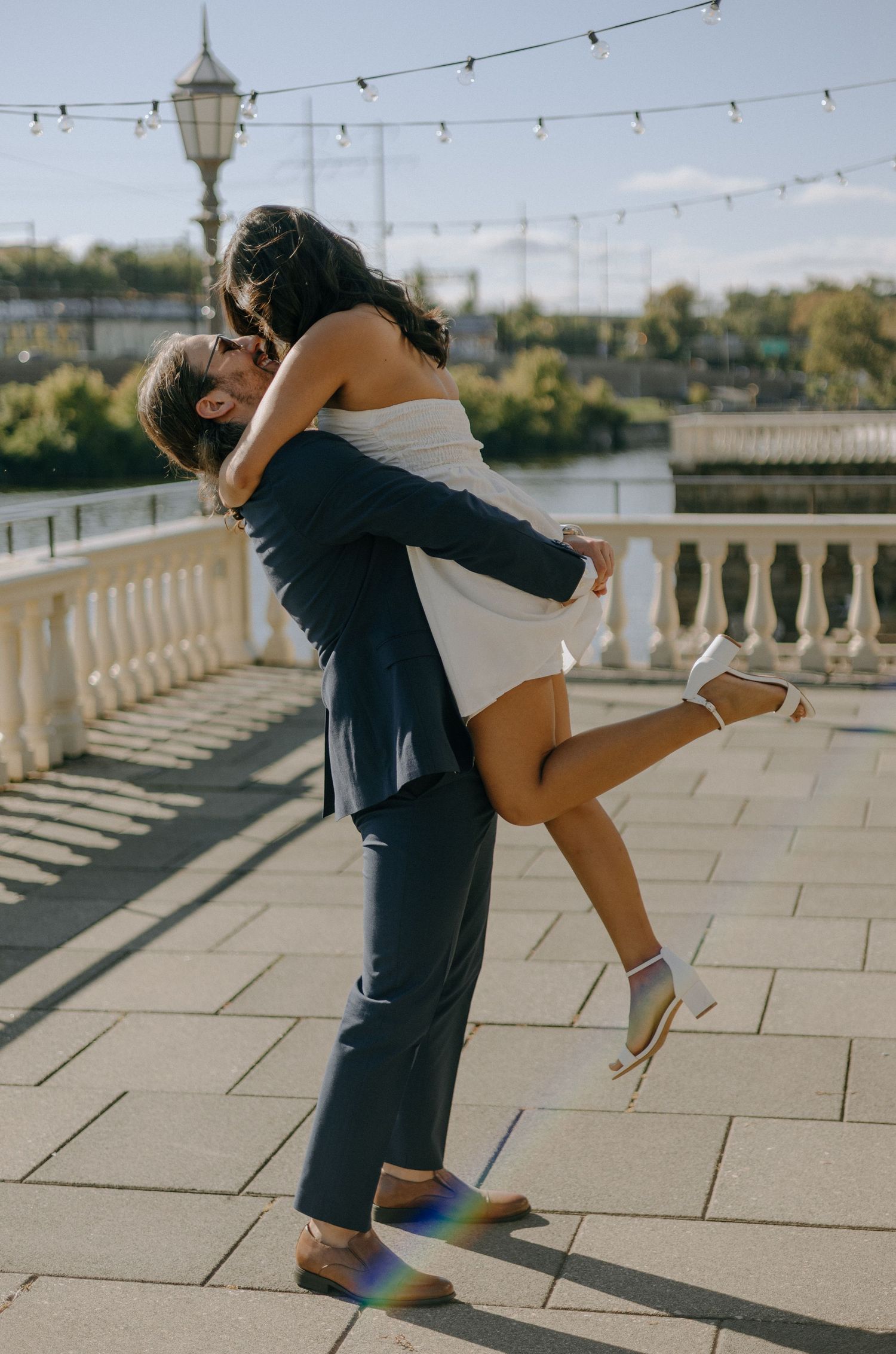 A couple dances and embraces on a sunlit terrace with string lights overhead and ornate stone balustrades.