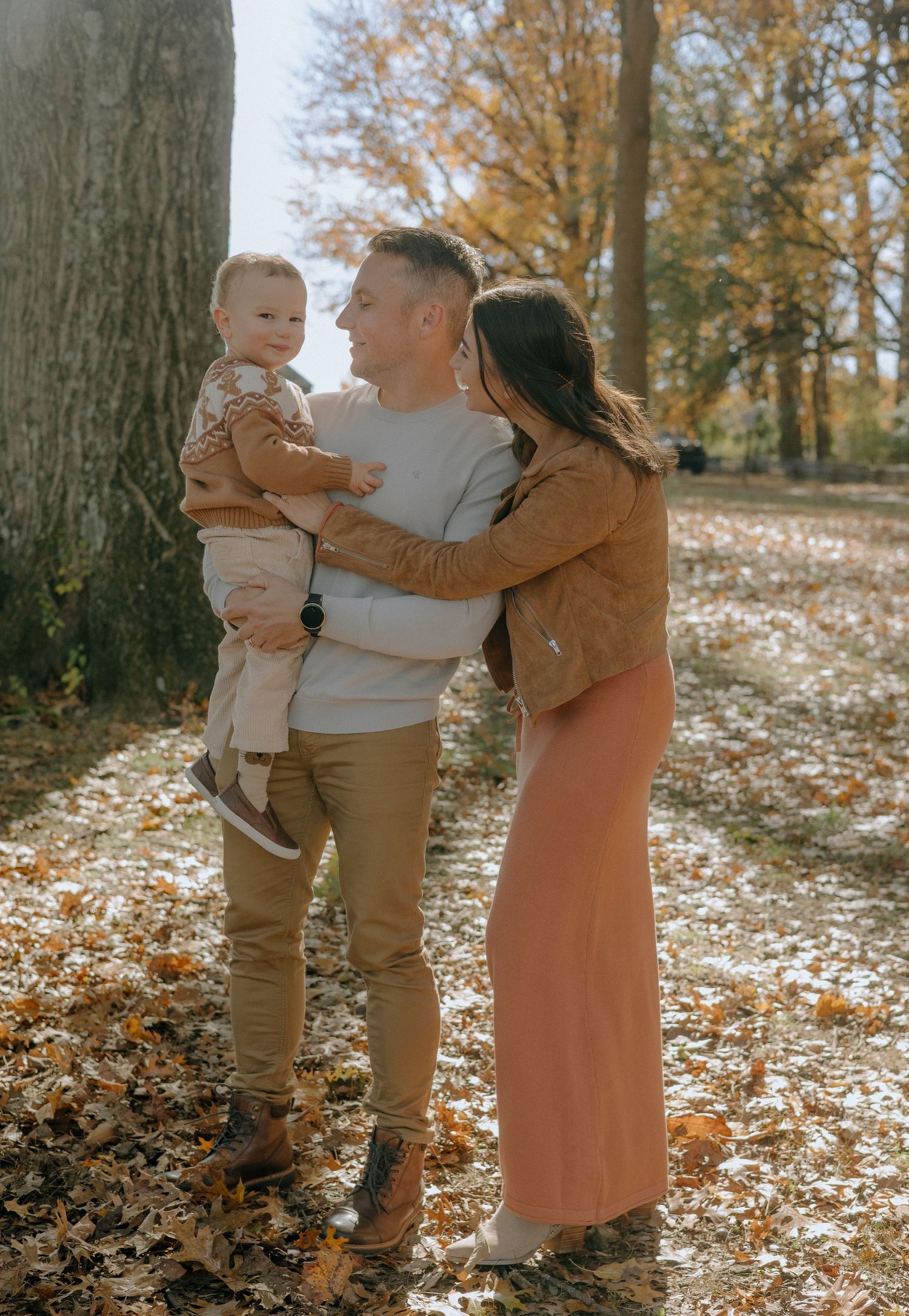 A family enjoys an autumn day together in a park with fallen leaves scattered on the ground.