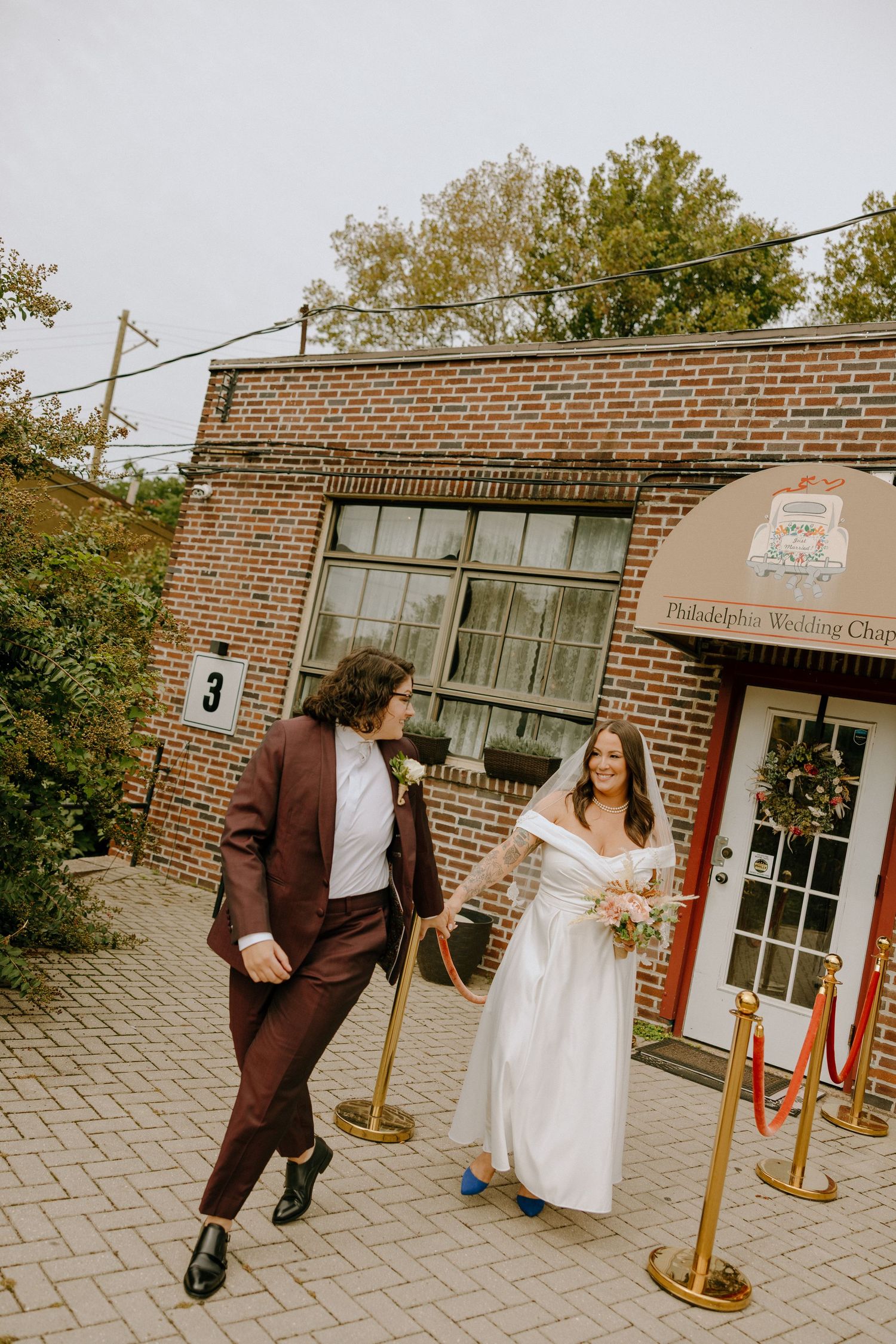 A couple in wedding attire playfully poses outside a brick building with stanchions and a storefront.