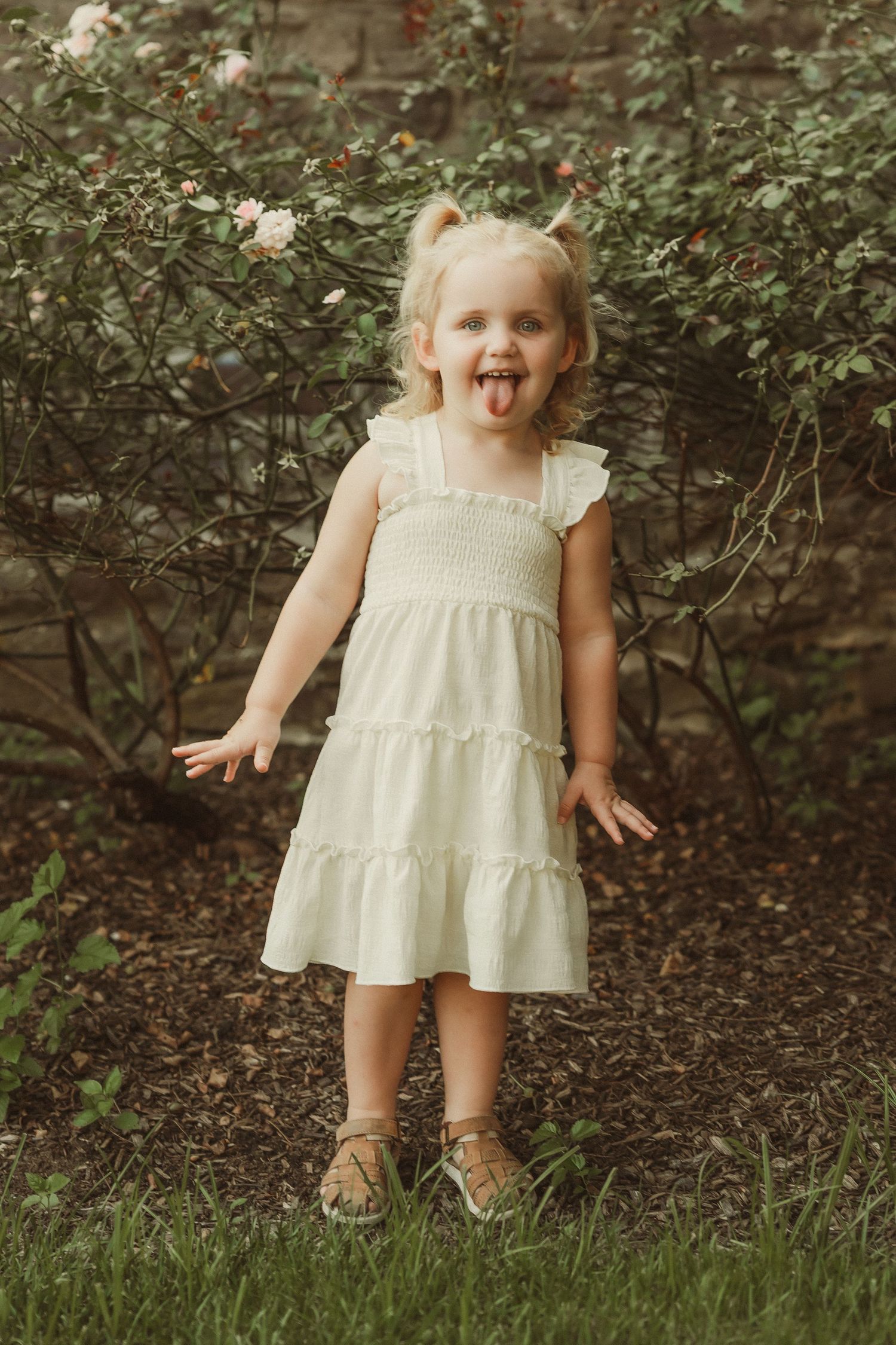 Person in white ruffled dress plays in garden setting near stone wall and bushes.