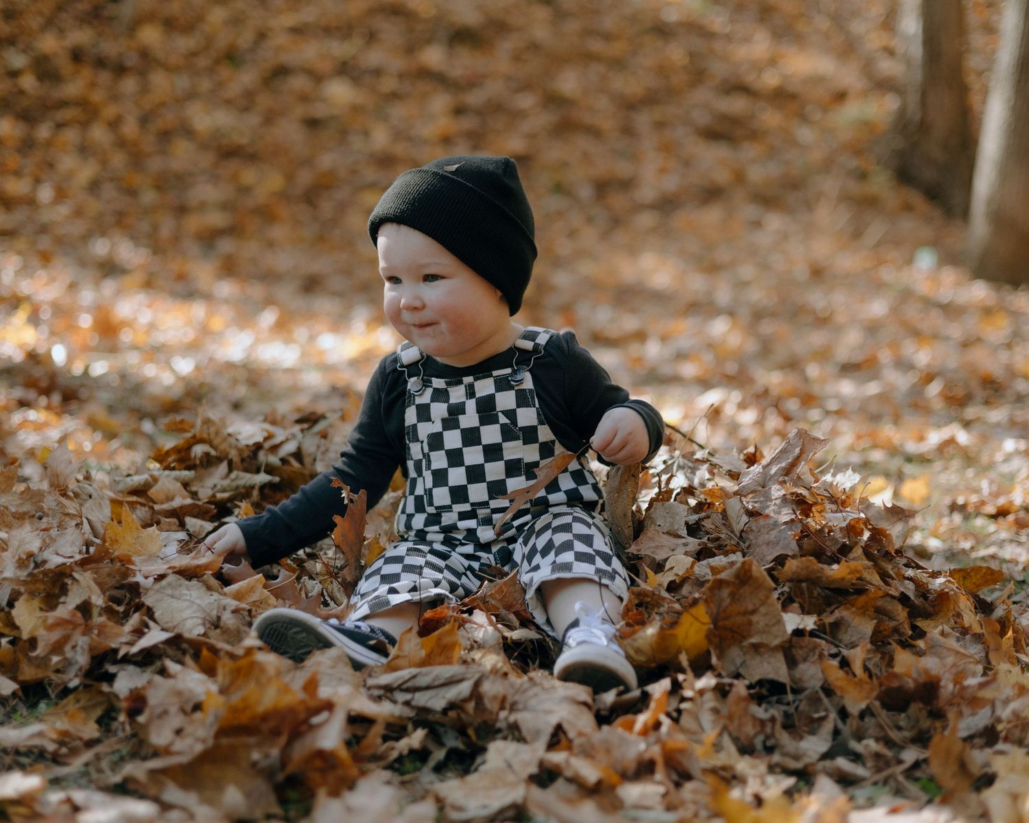 A small figure in a black beanie and checkered outfit plays in fallen autumn leaves on the ground.