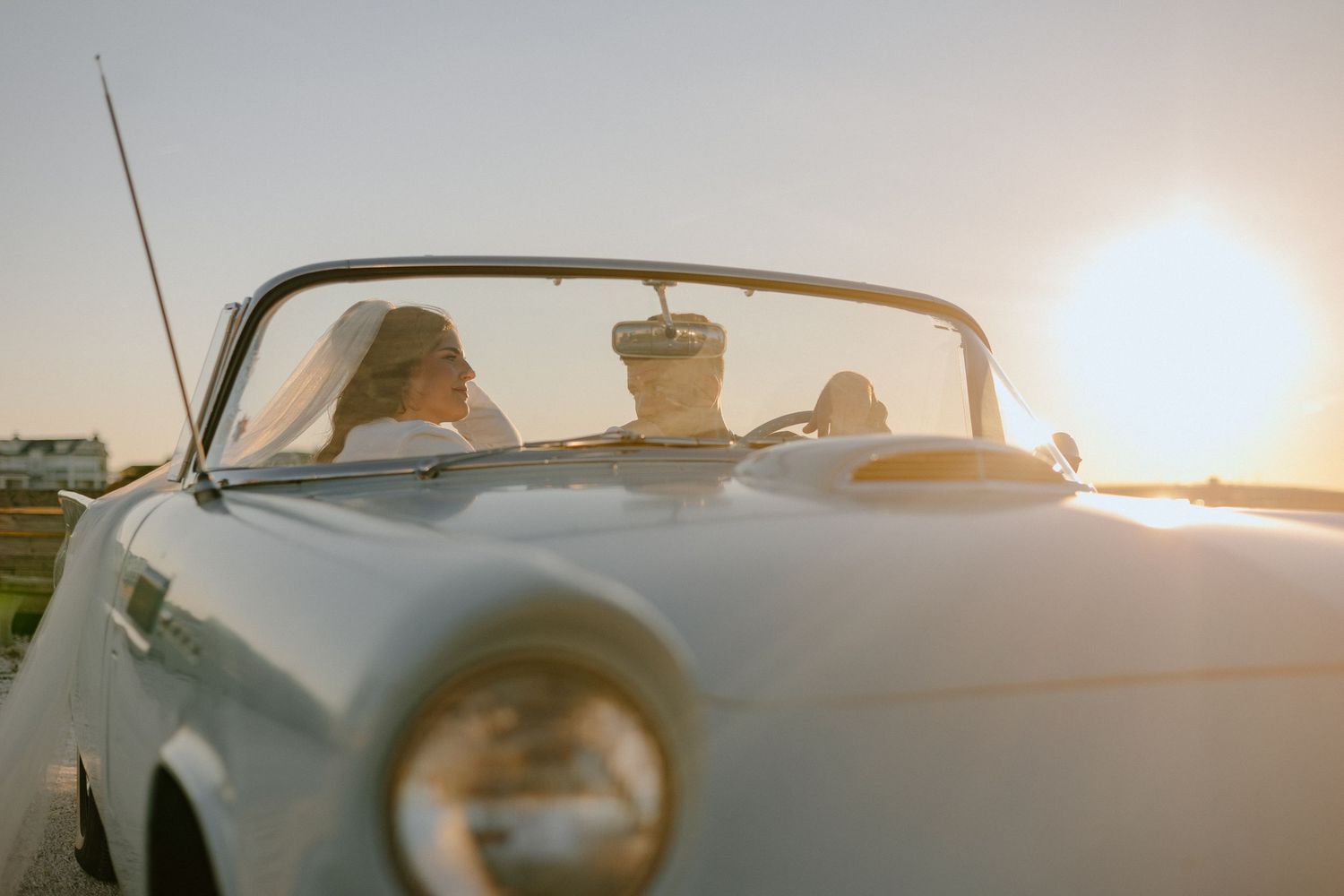 Classic convertible car silhouetted against golden sunset with sunlight flare and passengers enjoying scenic drive.