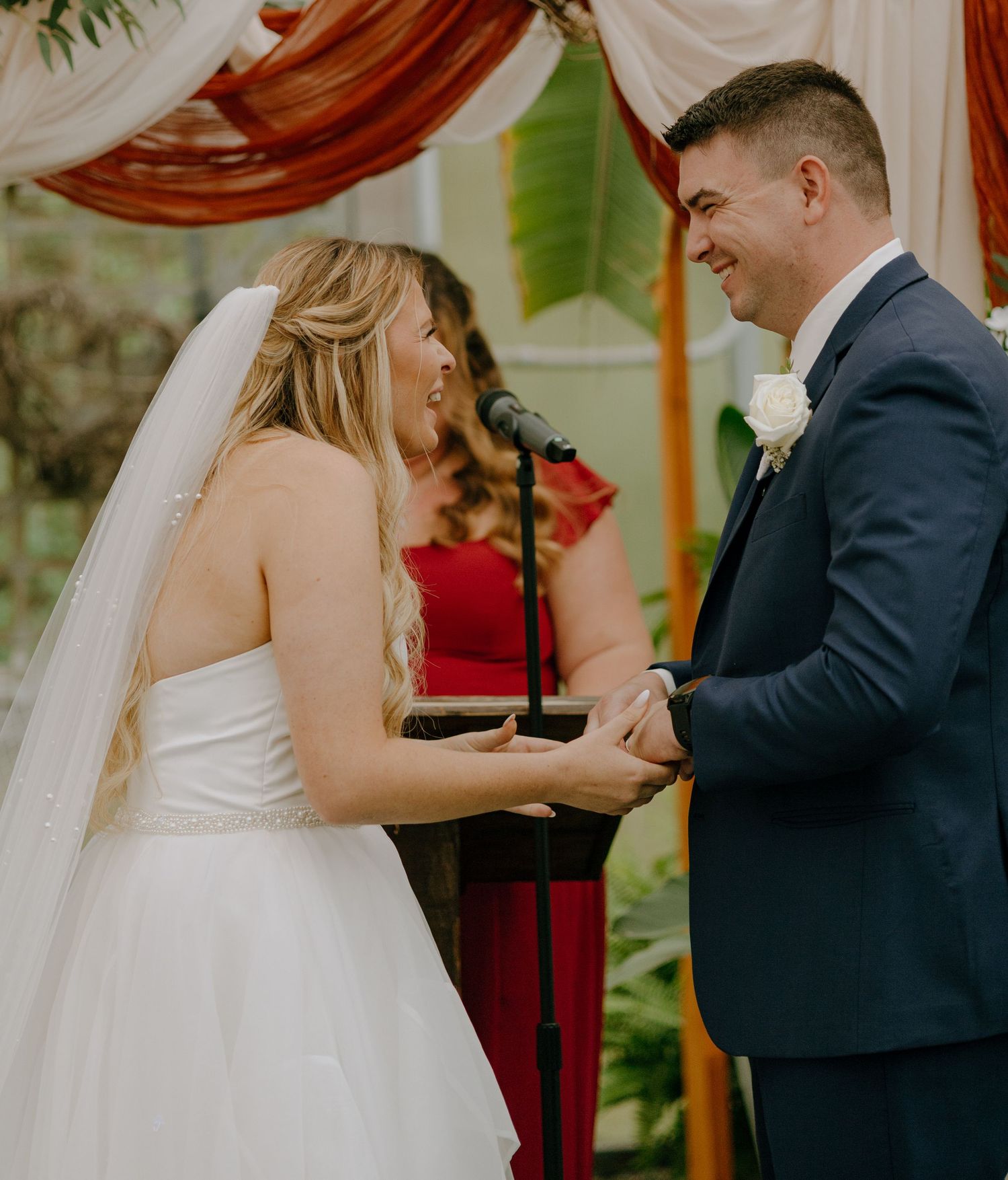 A couple exchanges vows during an outdoor wedding ceremony beneath draped fabric and greenery.