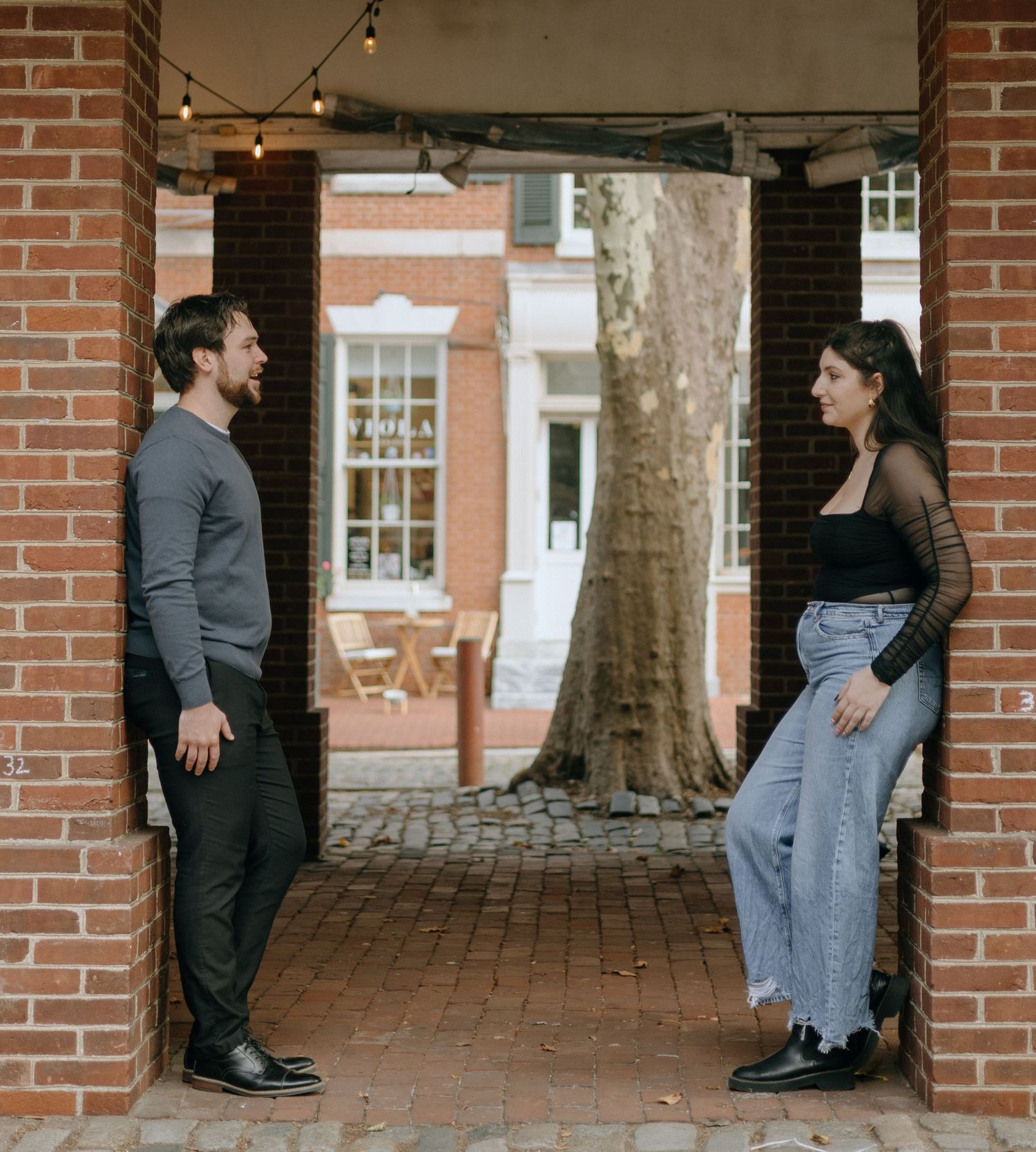 A couple stands together under a brick archway on a historic street with string lights overhead.