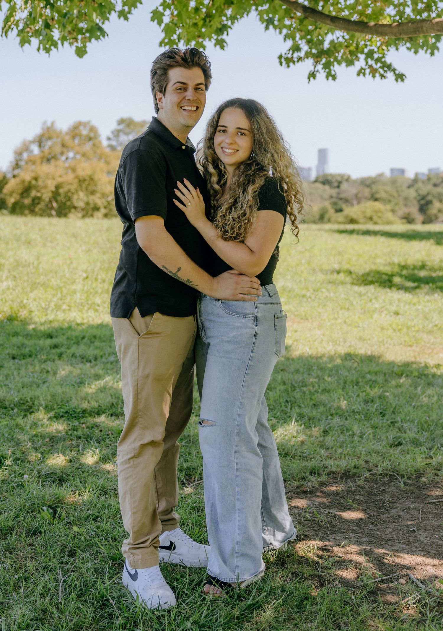 Romantic embrace between couple standing in grassy field with trees in background.