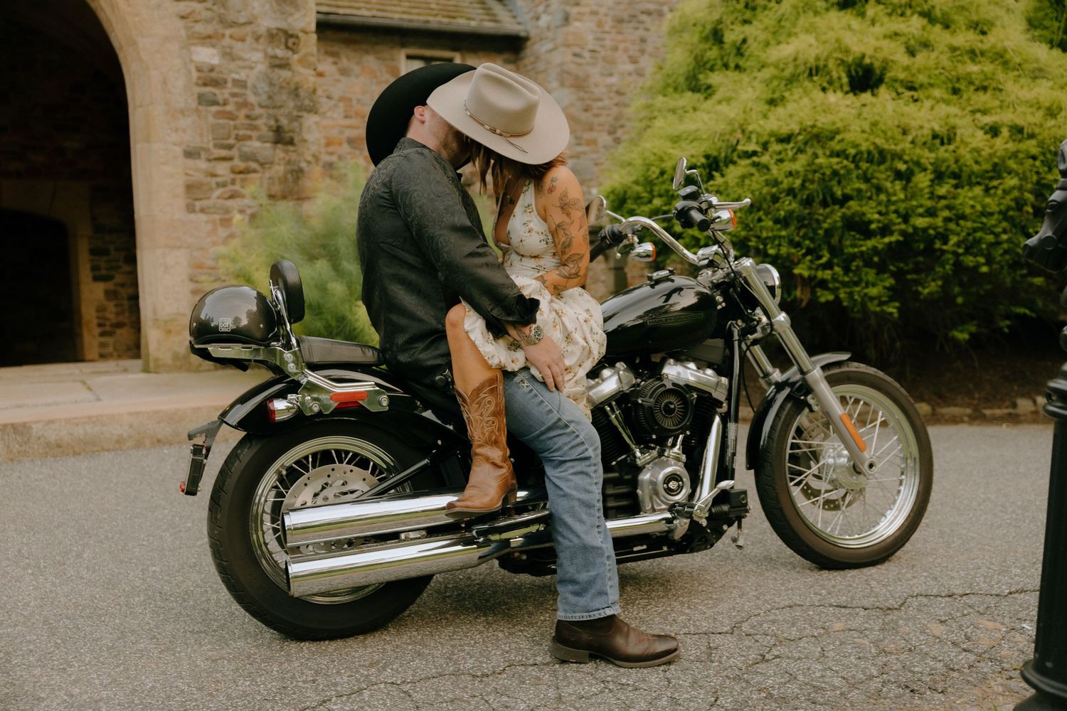 Couple poses casually near parked motorcycle outside stone building.