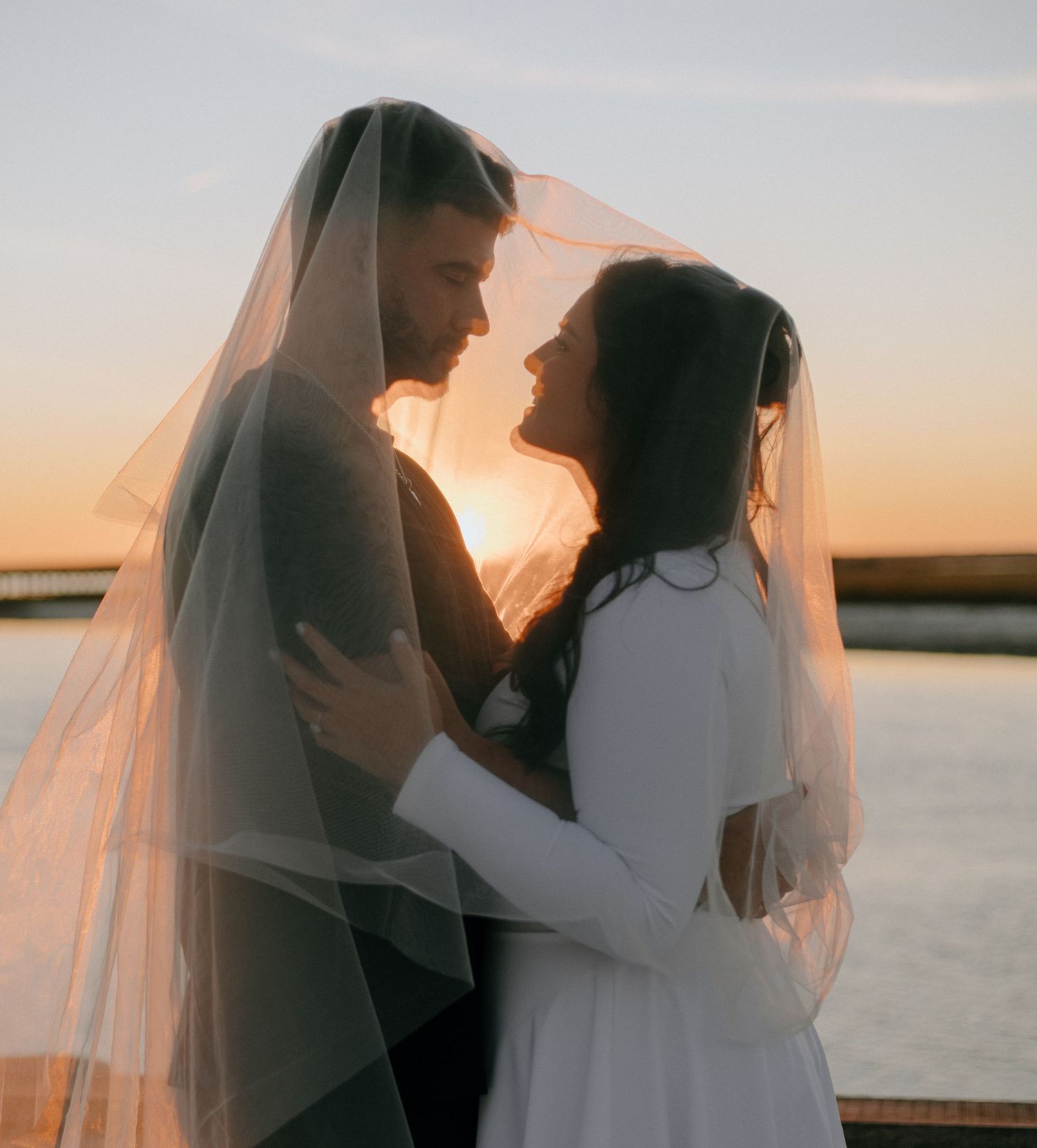 A romantic sunset silhouette of a couple in wedding attire embracing by the water's edge.