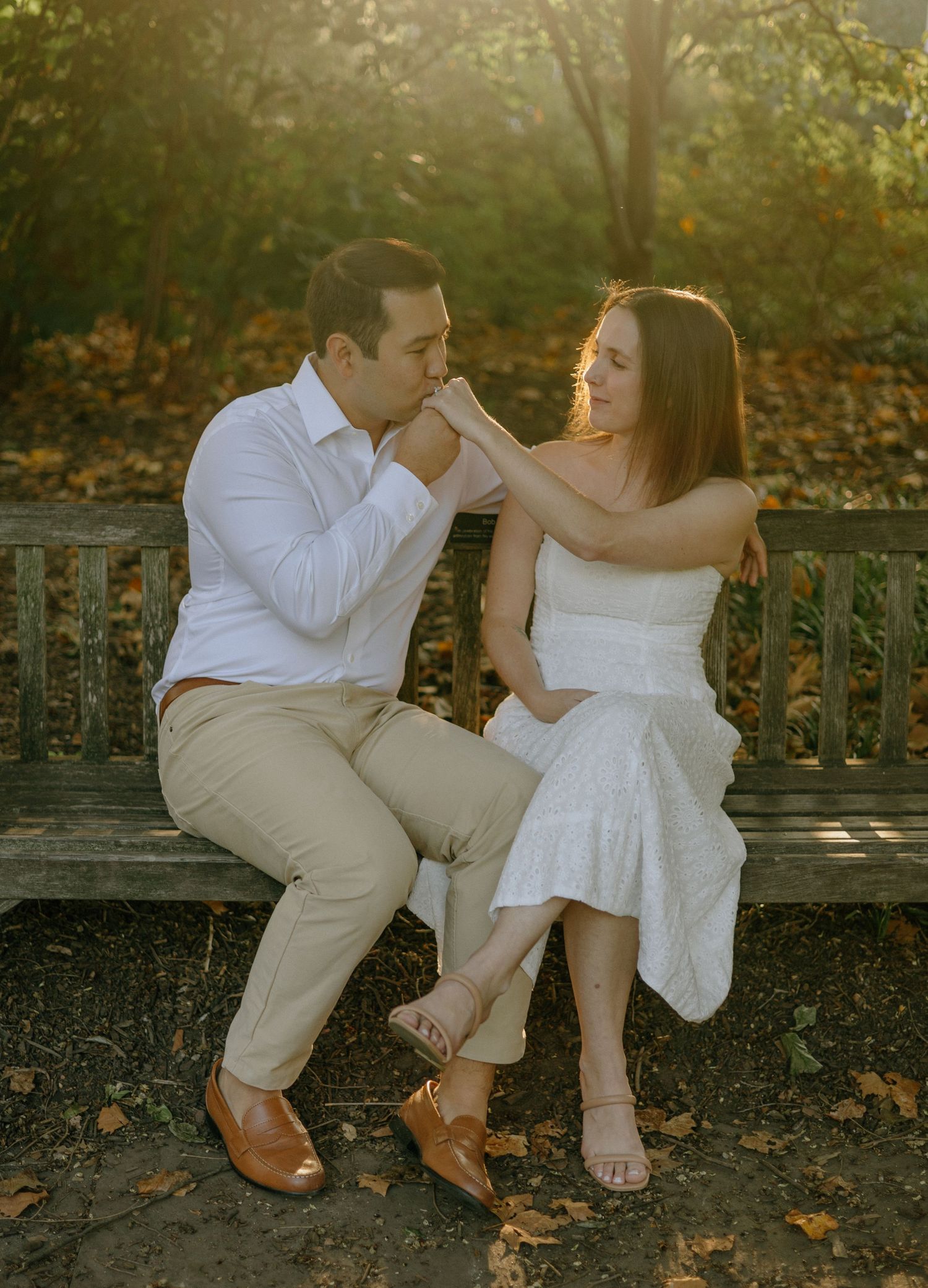 A romantic couple shares intimate moments while sitting on a stone bench surrounded by autumn foliage at sunset.