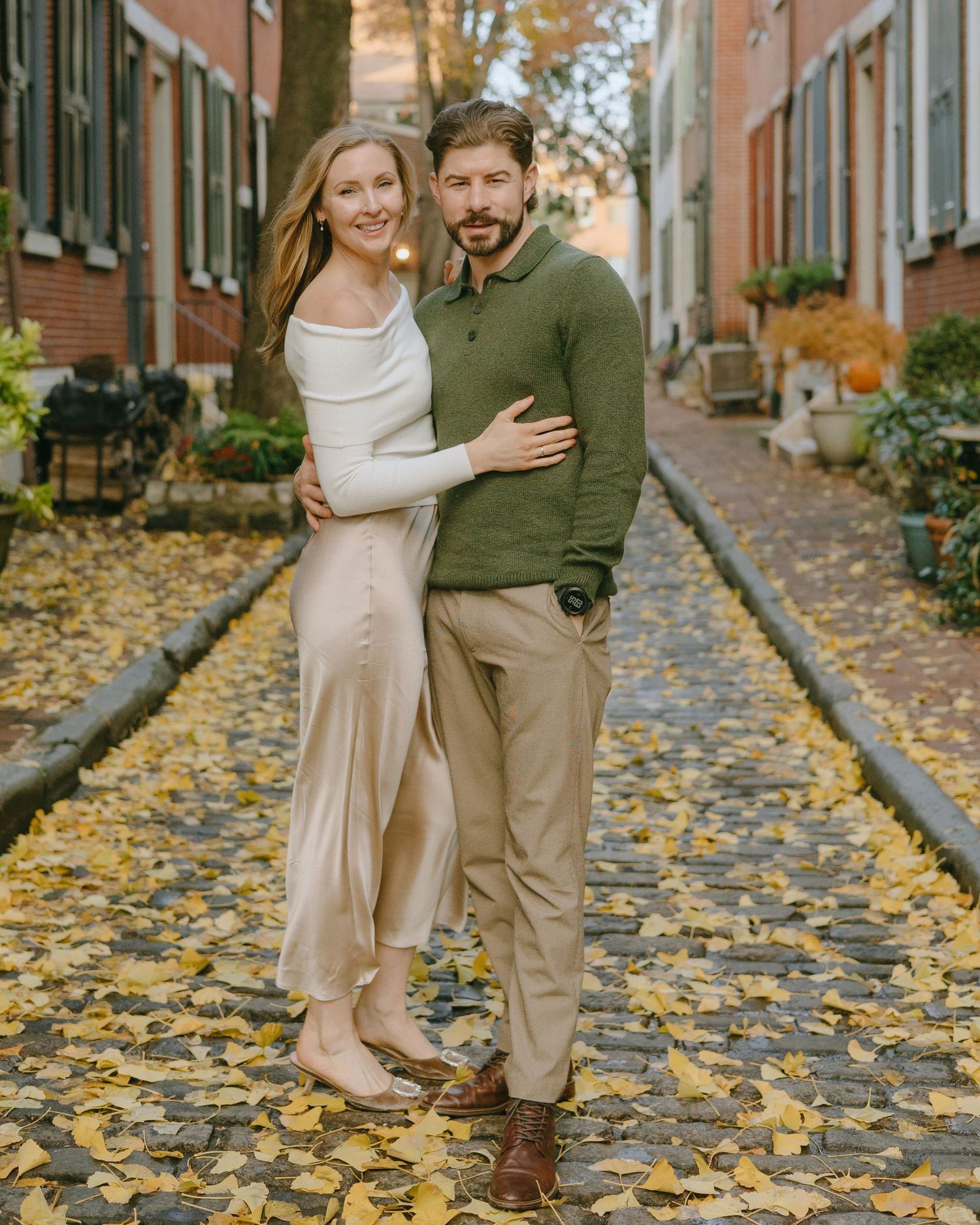 A couple celebrates on a charming cobblestone street covered in yellow autumn leaves in a historic neighborhood.
