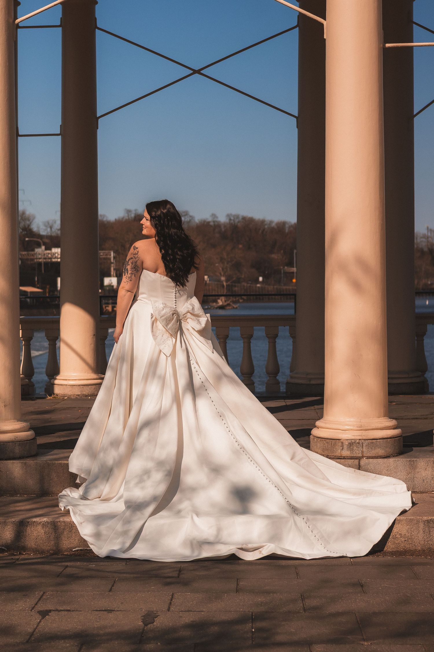 Elegant white wedding dress with long train photographed between classical stone columns near water.