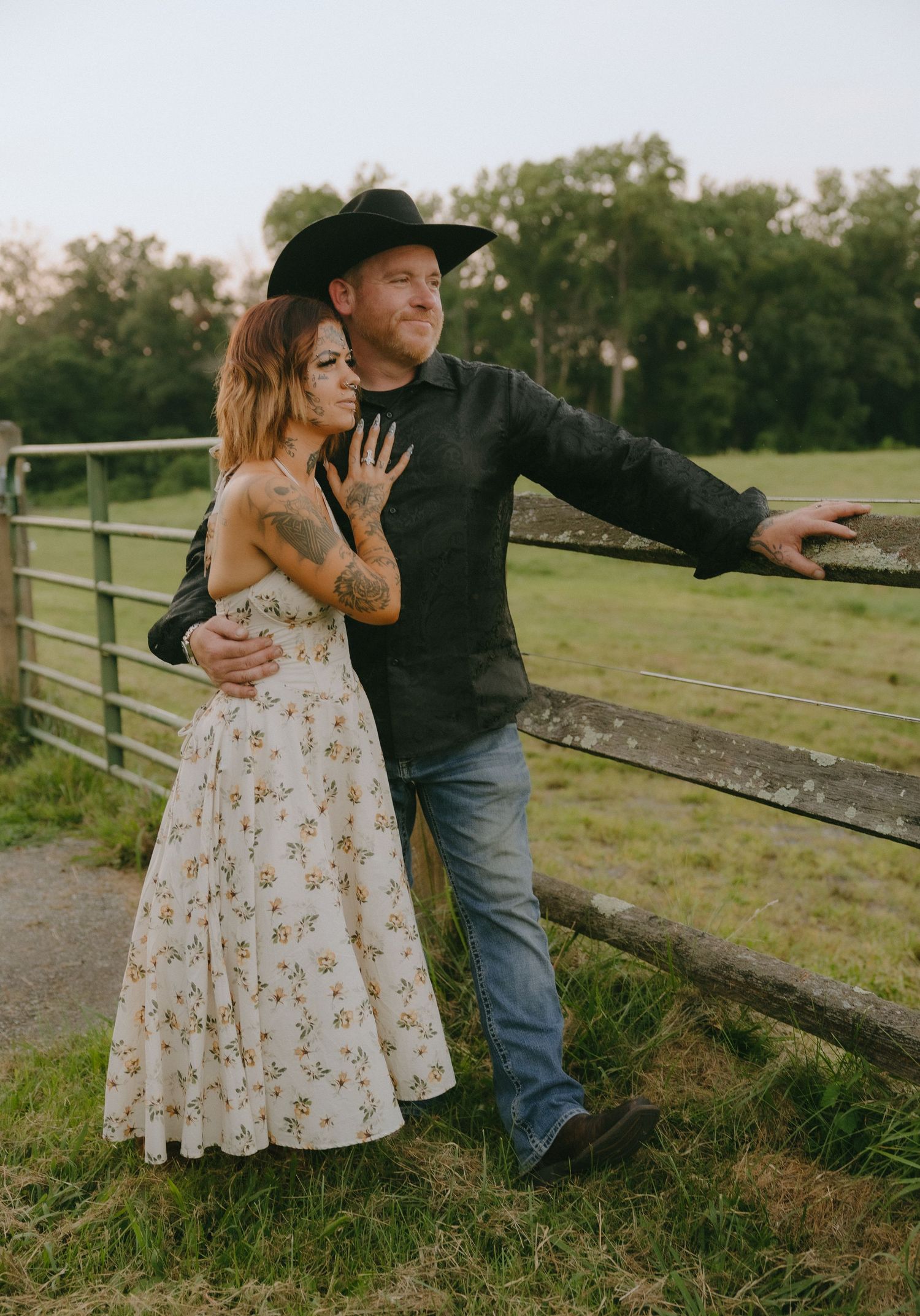 A couple shares a romantic moment leaning against a rustic wooden fence in a pastoral country setting.