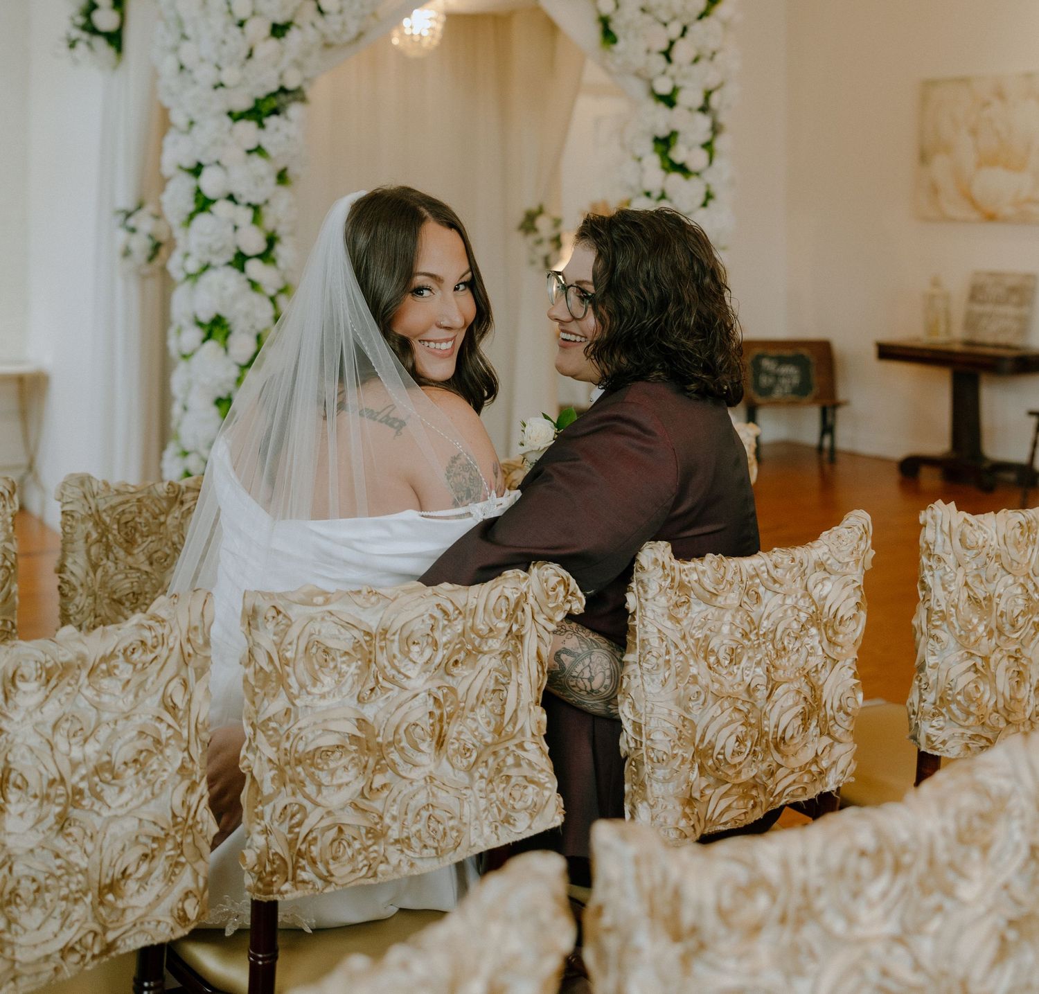 A wedding couple shares an intimate moment while seated in ornate chairs under a floral arch.