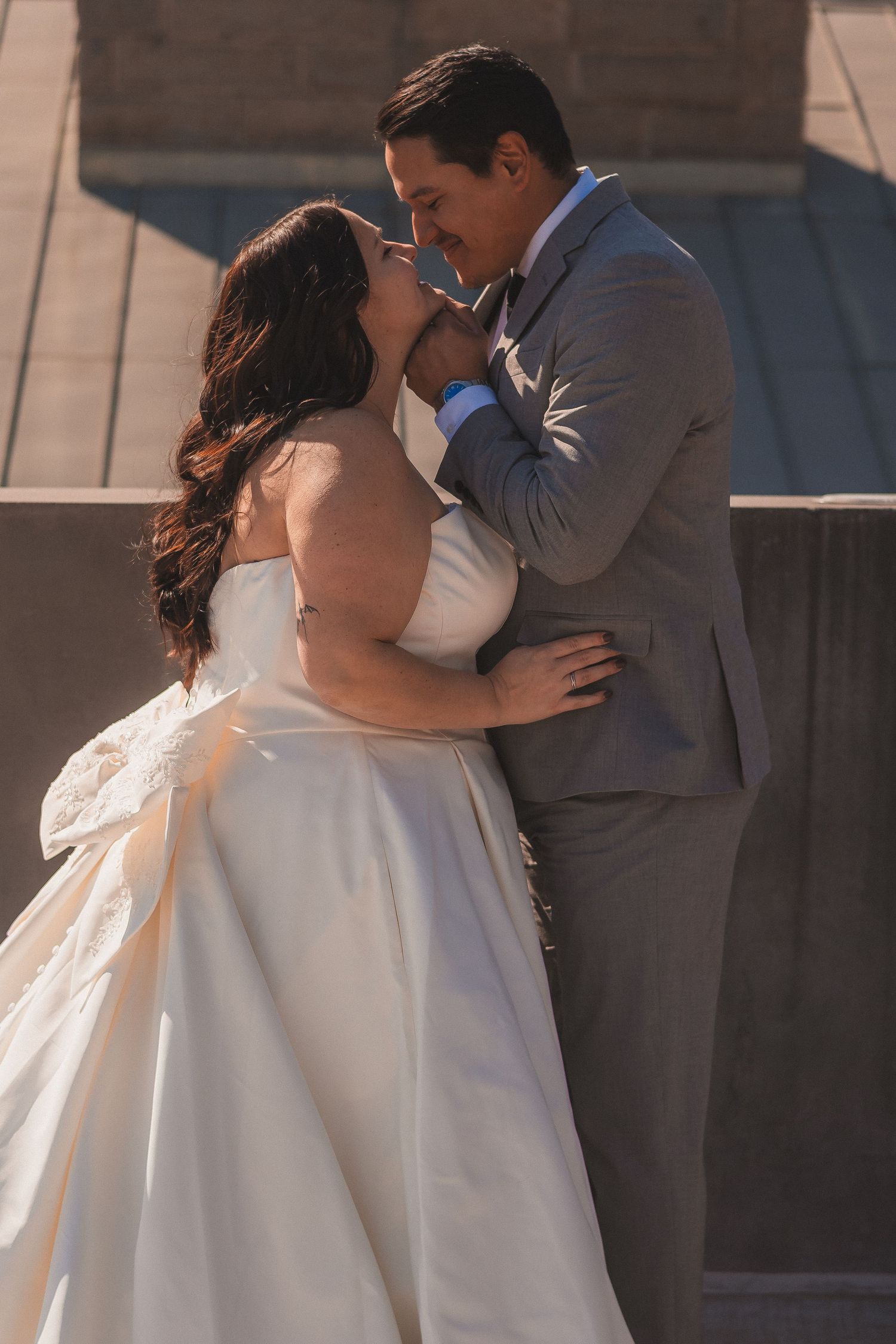 Romantic wedding couple in white dress and grey suit embrace in tender moment during sunset photoshoot.