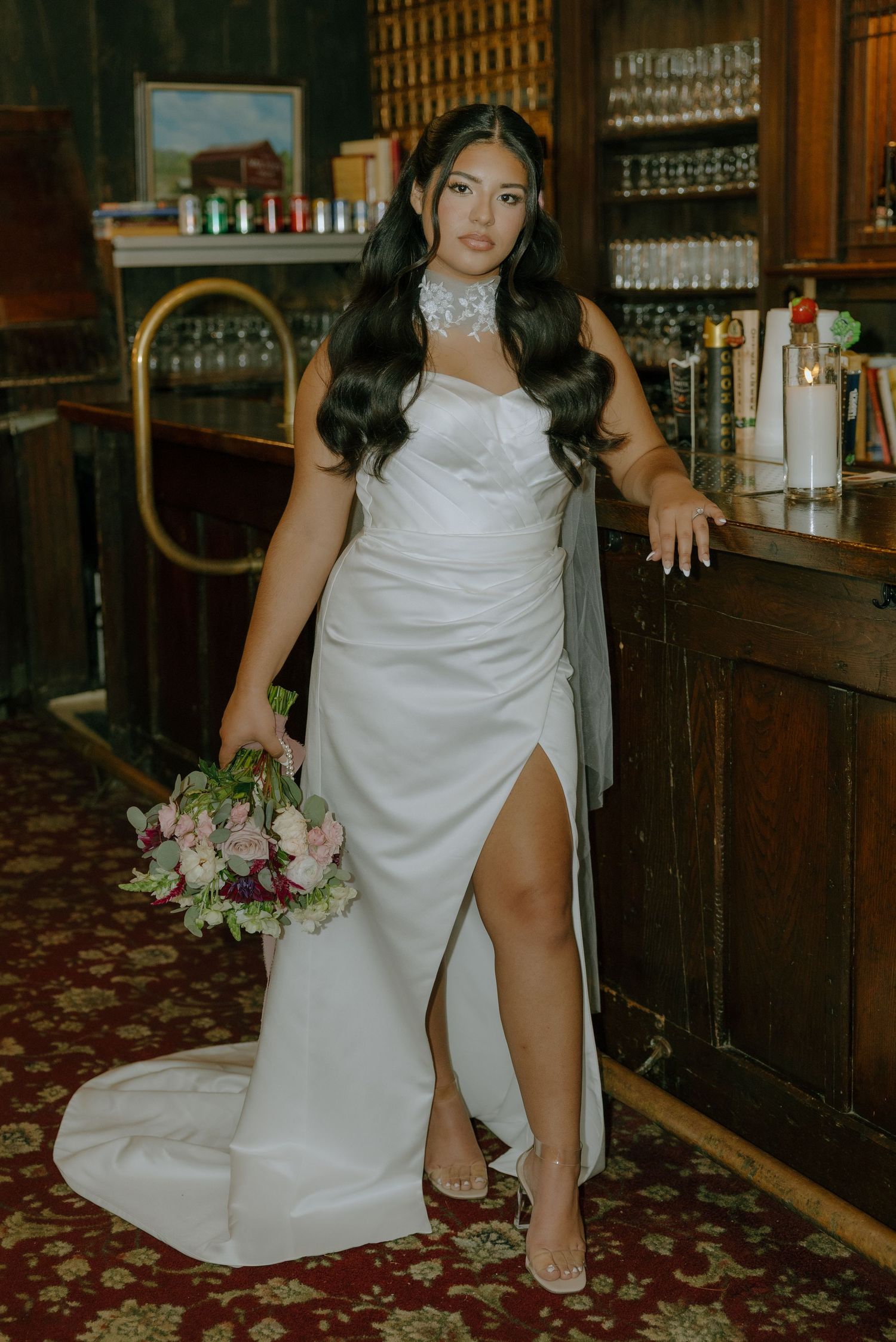 A bride in a white wedding dress poses with a bouquet in front of a vintage wooden bar.