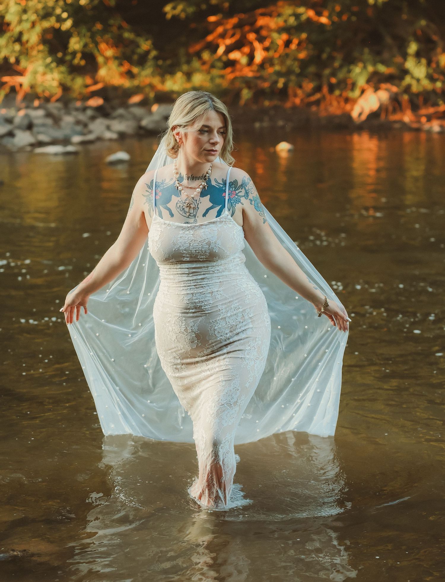 A bride wearing a fitted lace dress stands in river water while holding her veil during sunset.
