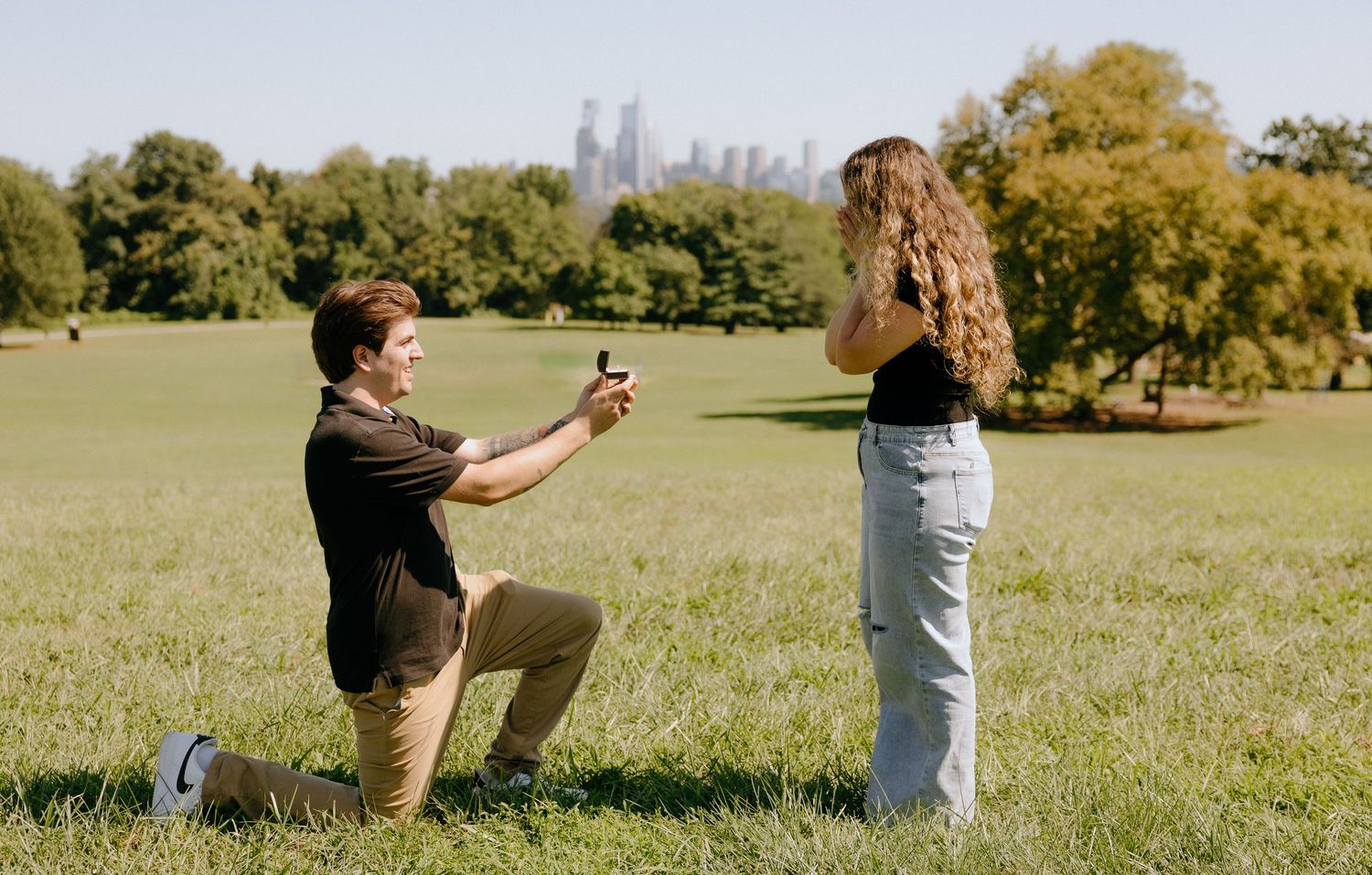 Proposal moment captured in sunny park with city skyline visible in background.