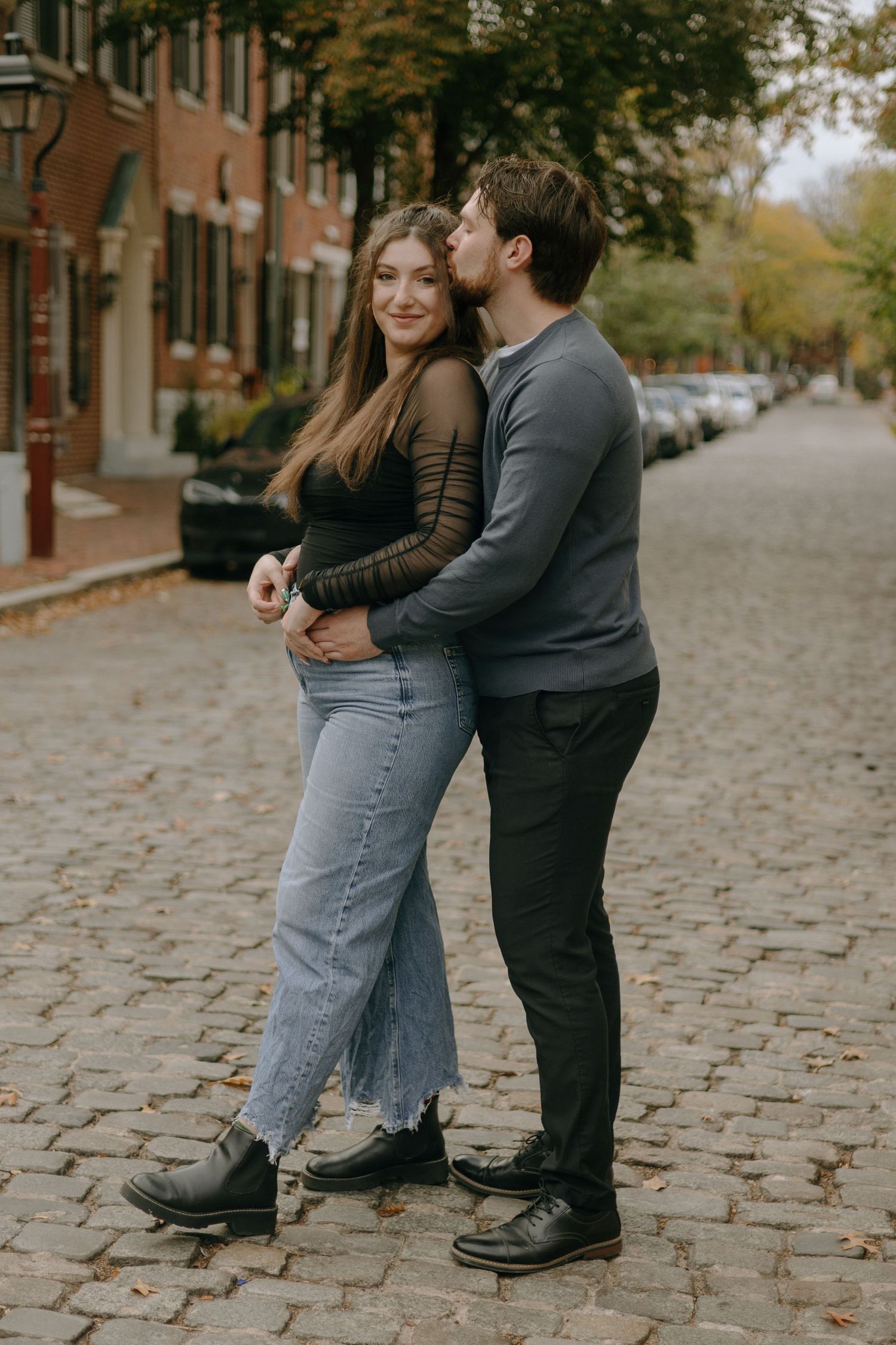 A couple shares a romantic moment on a cobblestone street lined with brick buildings during autumn.