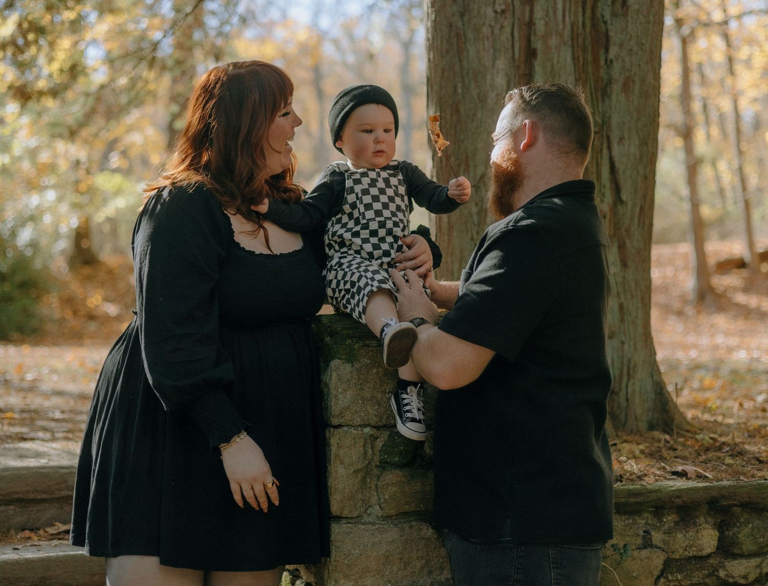 Two people in black outfits share an intimate moment in an autumn forest setting with fallen leaves and trees.