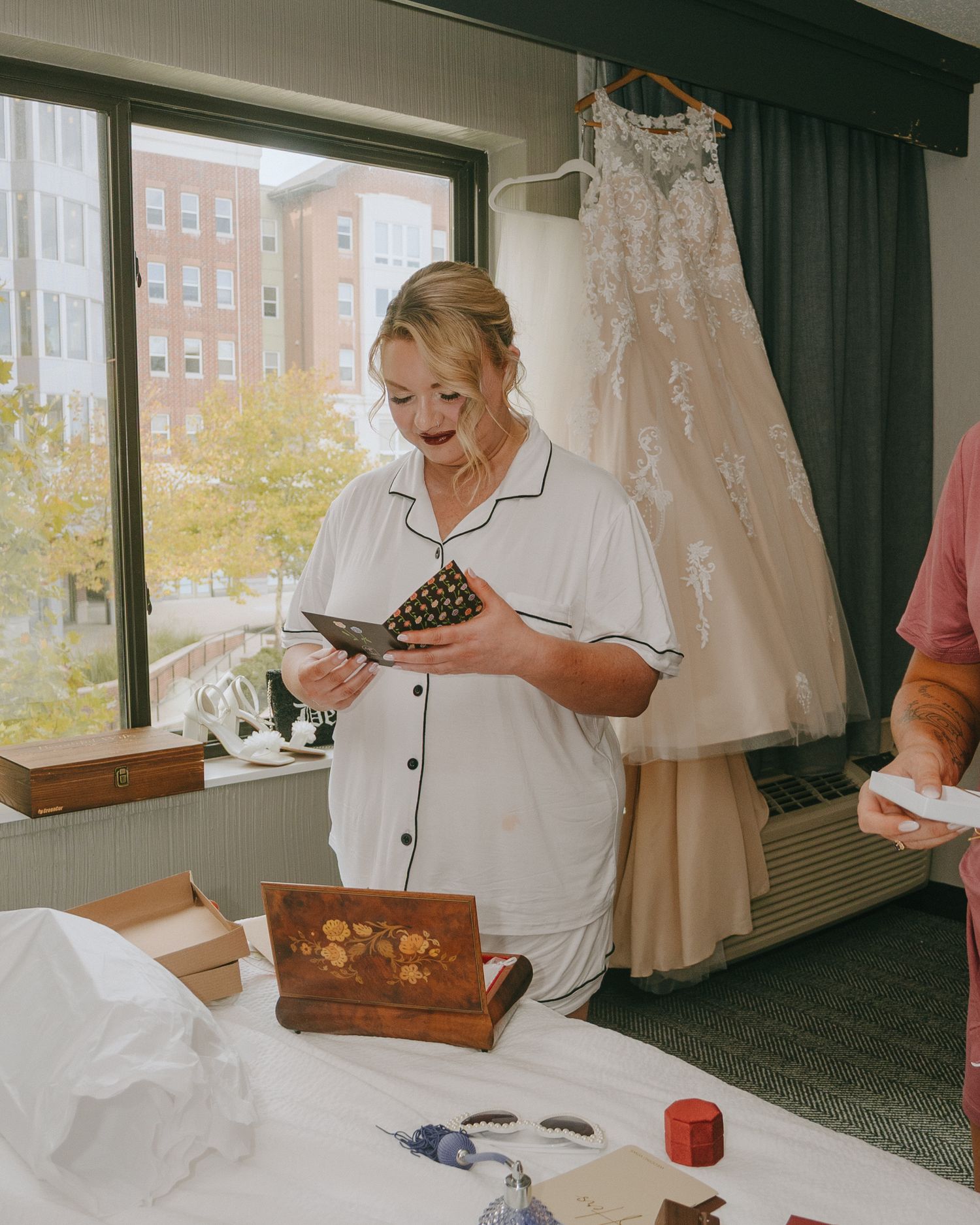 Wedding preparations show a white lace dress hanging while someone opens a decorative box.