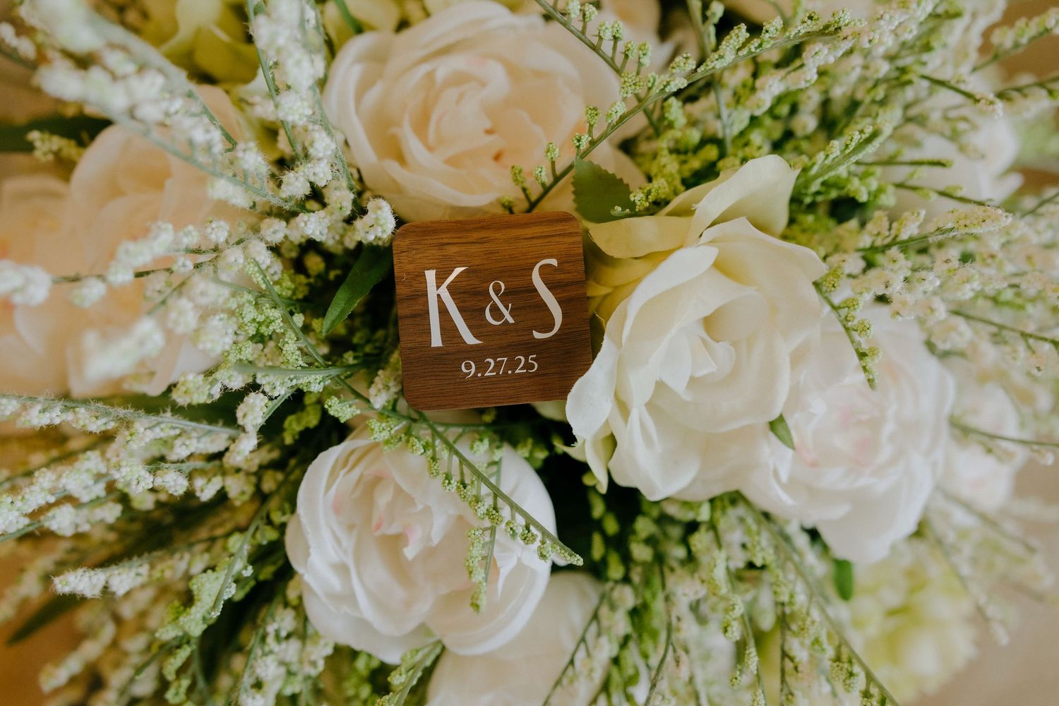 Close-up of a wedding bouquet with white roses and greenery featuring a wooden K&S date marker.