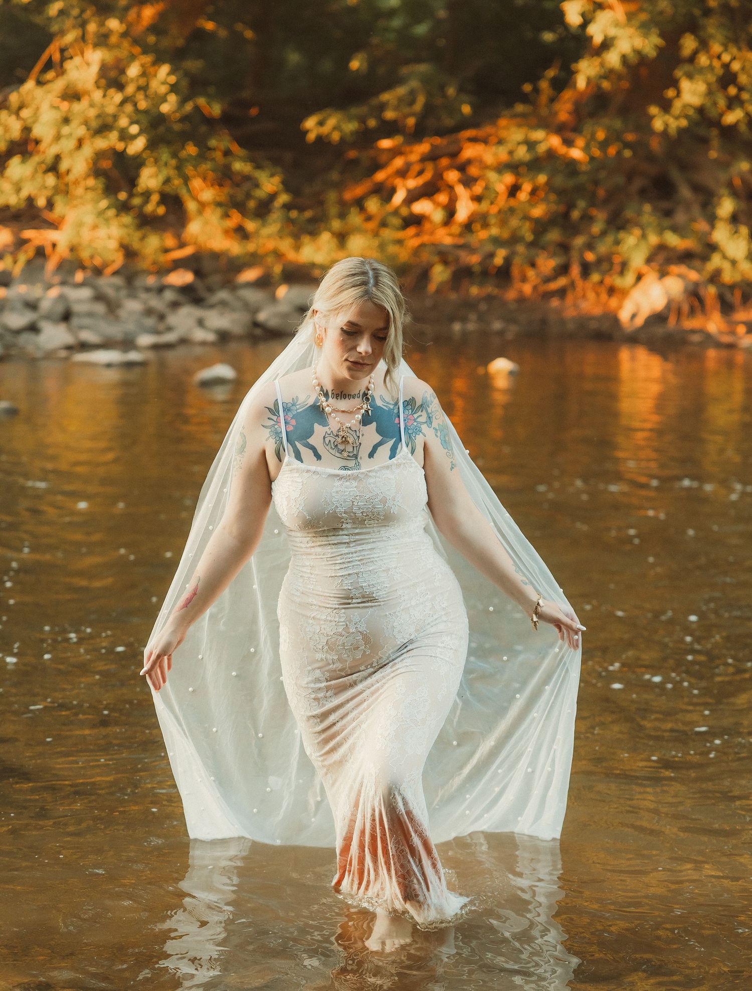 A bride wades through river water in a fitted white dress with blue floral details and flowing veil.